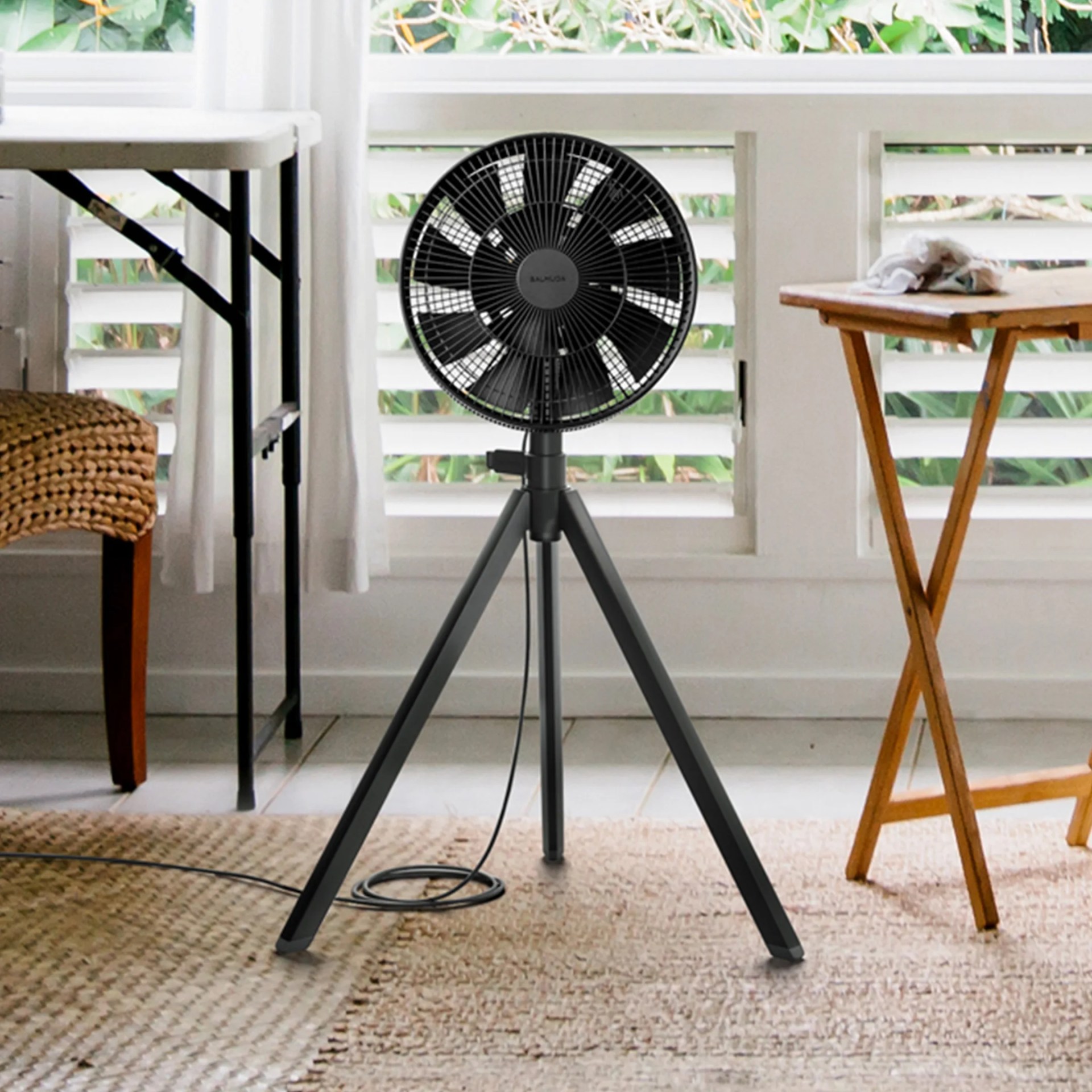 Black tripod floor fan with circular grill and visible power cord on a woven rug near wooden and metal tables.