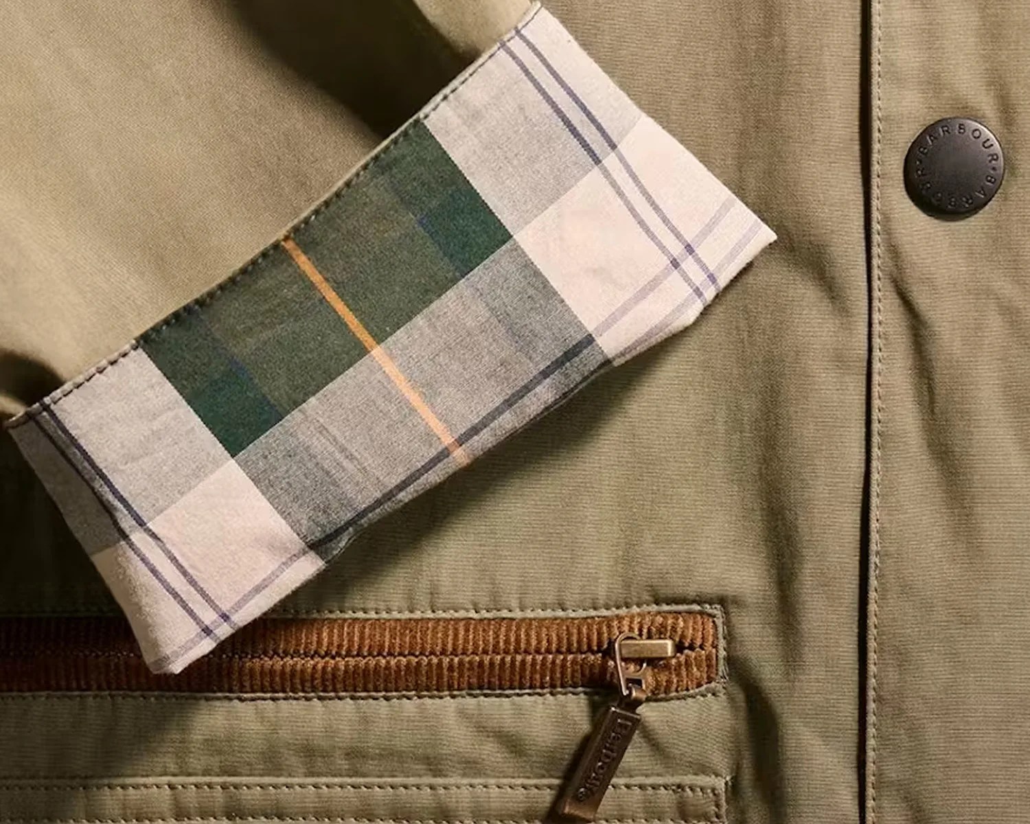 Close-up of a khaki jacket sleeve with a plaid cuff, a brown corduroy-trimmed pocket, and a black button with "Barbour" text.