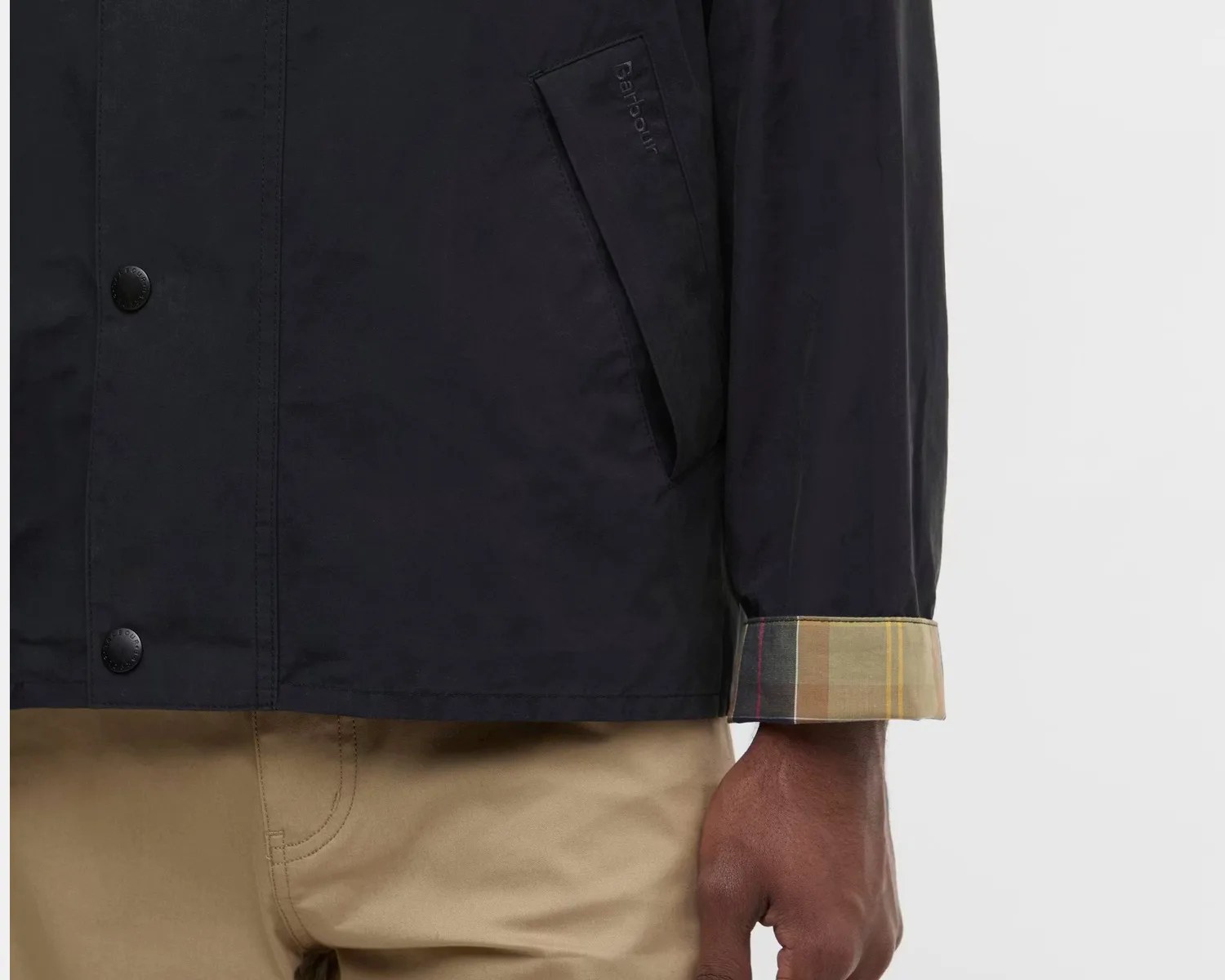 Black Barbour jacket with snap buttons and a plaid cuff worn with beige pants.