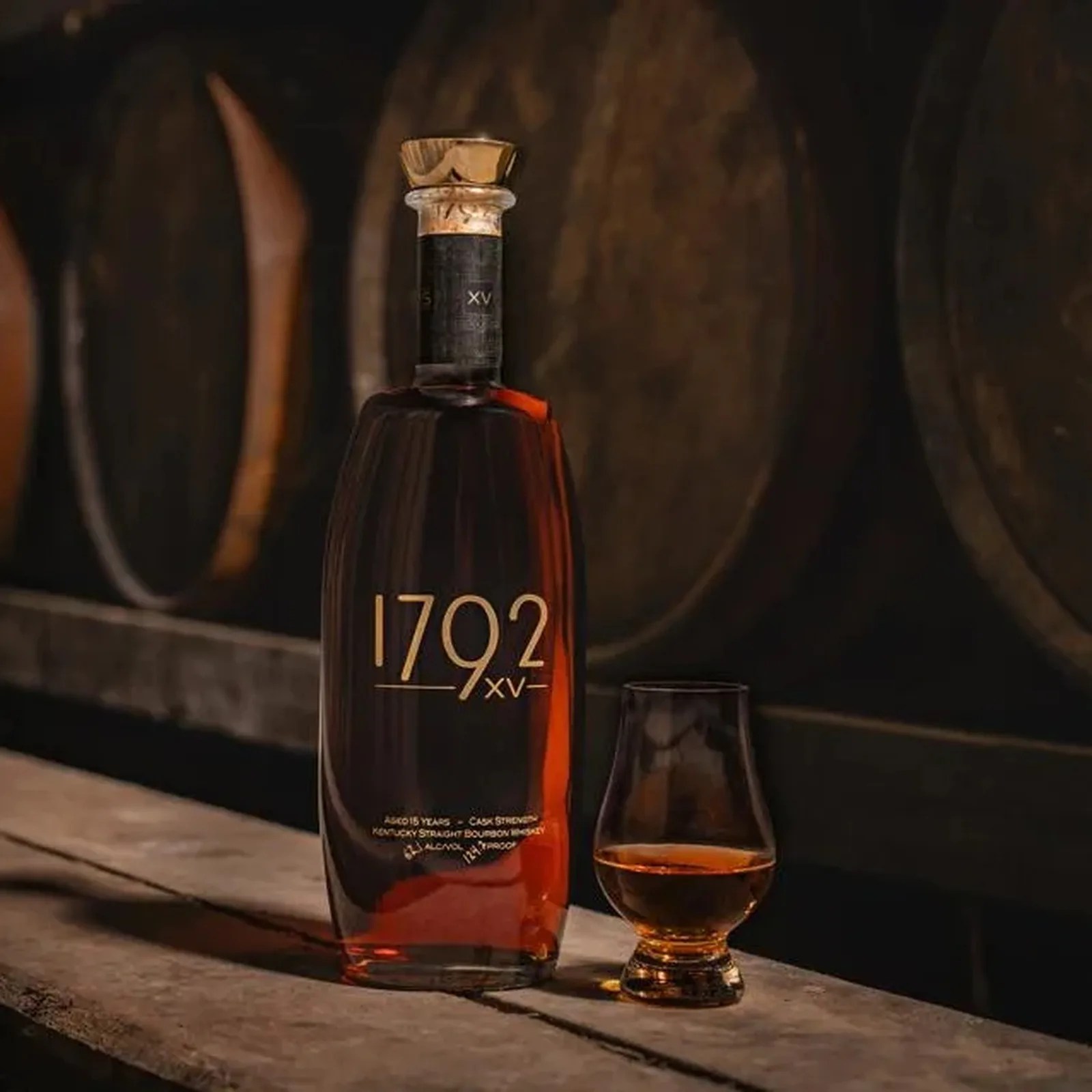Tall bottle of 1792 XV bourbon whiskey with a filled glass on a wooden surface in front of barrels.