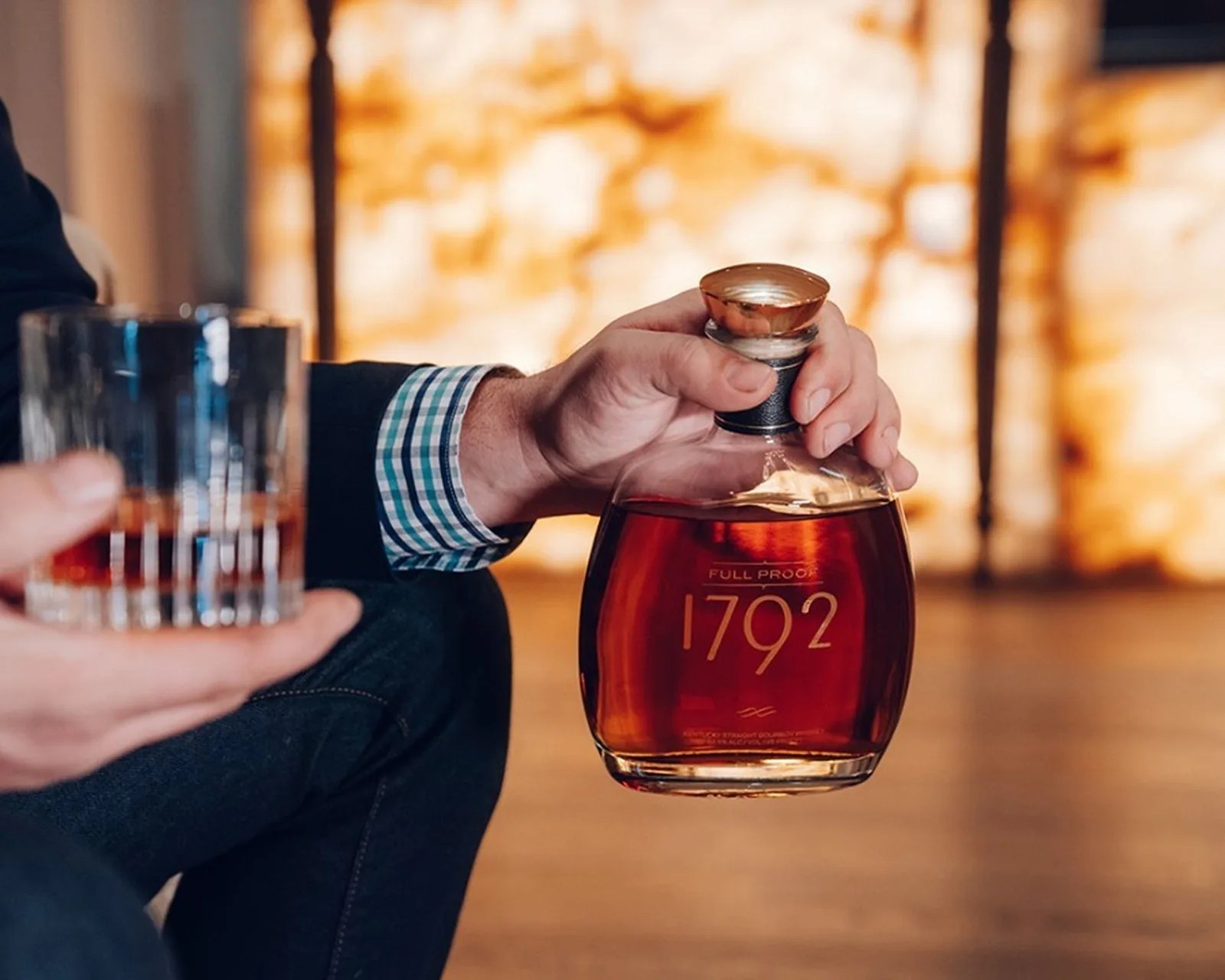 Hand holding a glass of whiskey and a bottle labeled "1792 Full Proof" with amber liquid inside.