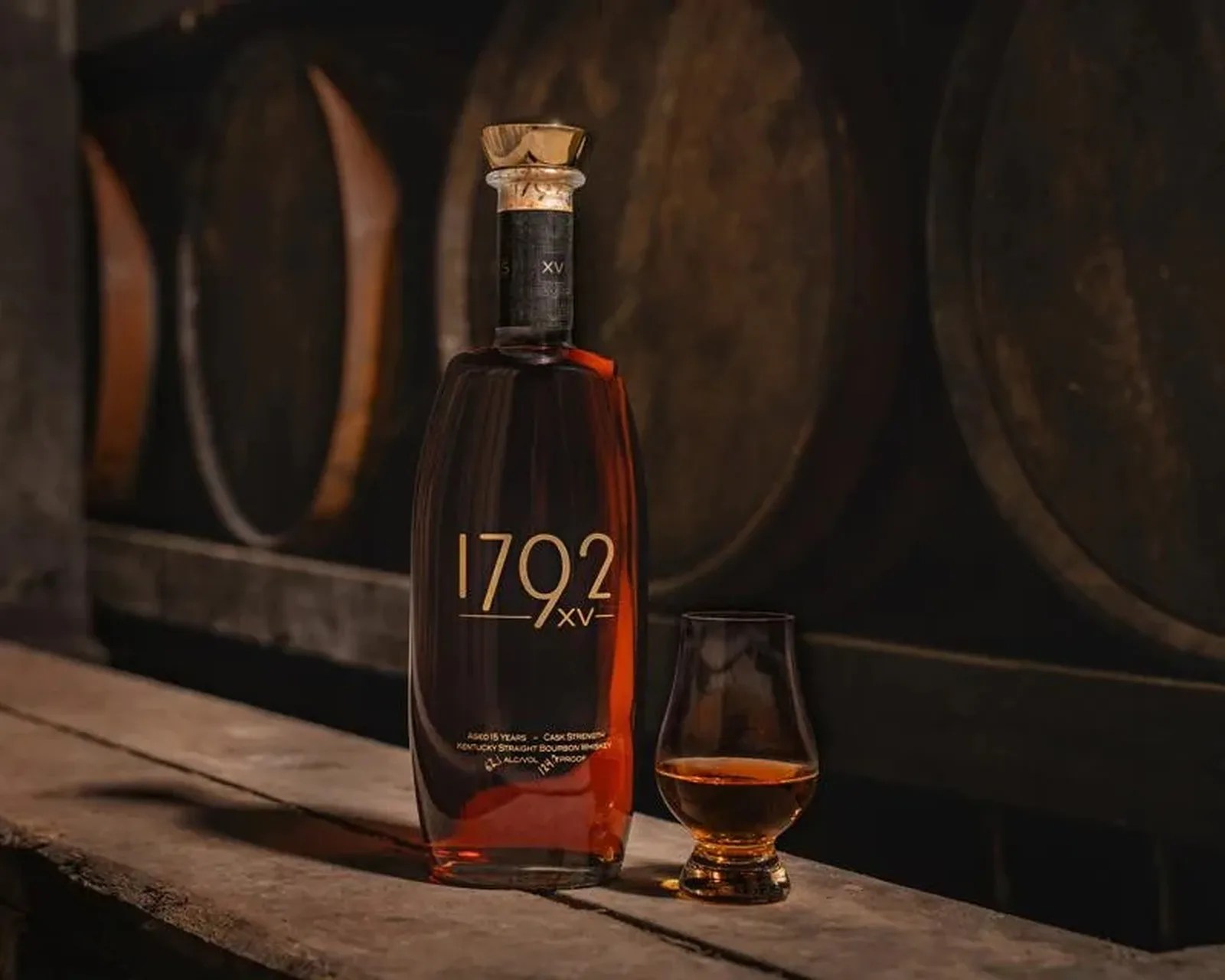 Bottle of 1792 XV bourbon whiskey and a filled glass on a wooden surface with barrels in the background.