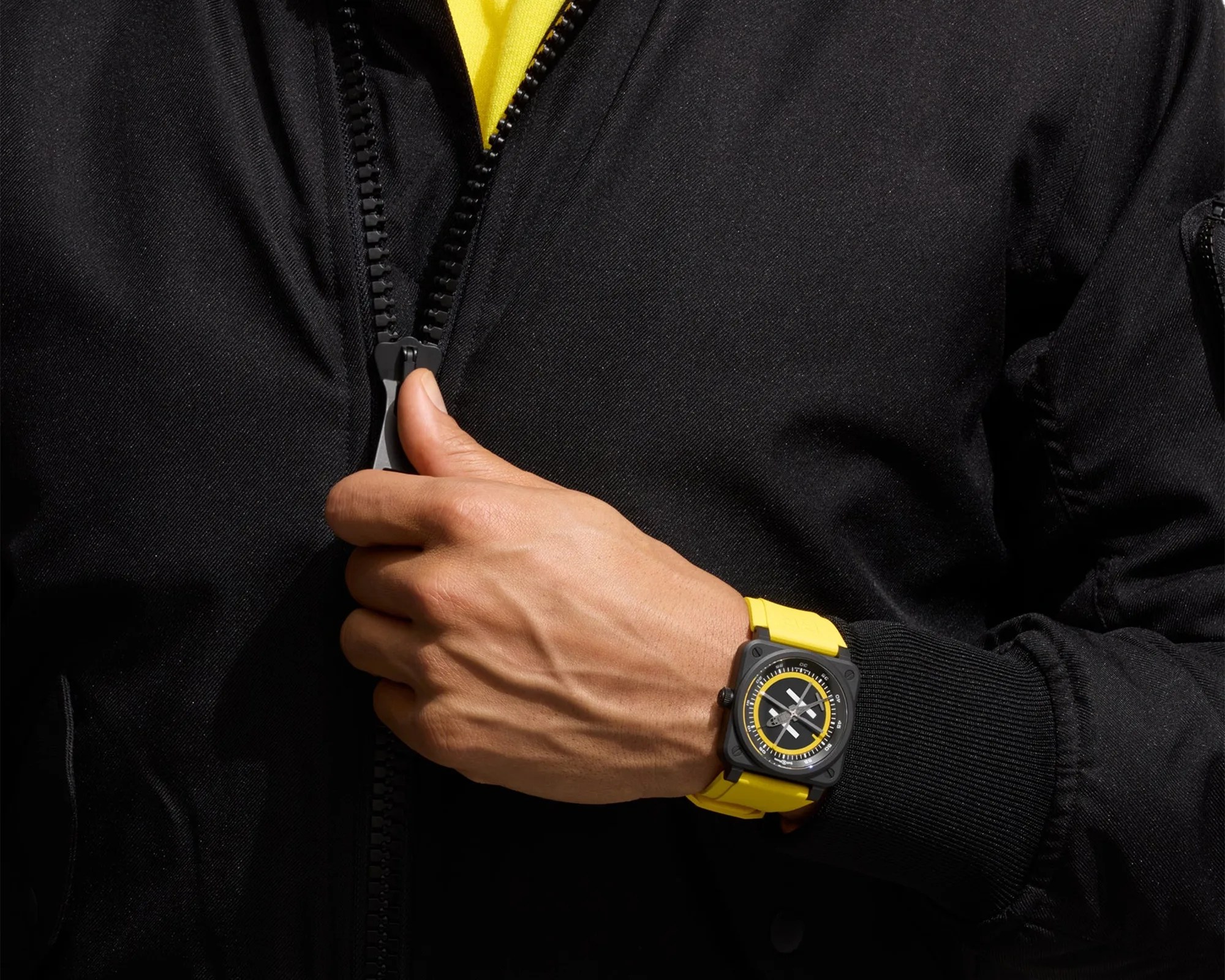 Hand zipping up a black jacket, wearing a black and yellow wristwatch with a square face.