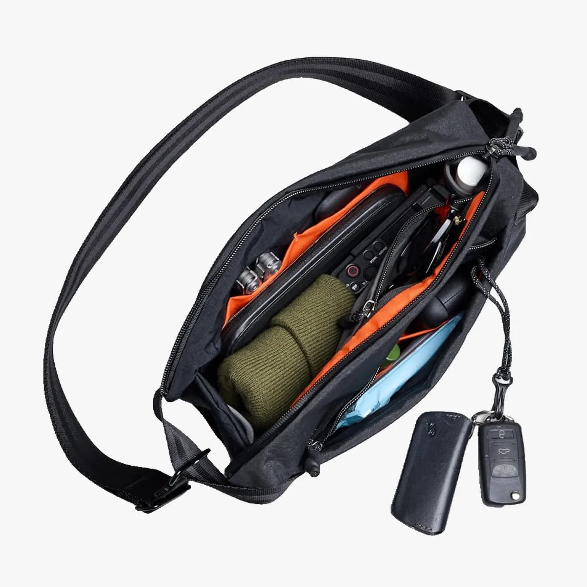 Black crossbody bag with orange interior pockets holding a green knit item, sunglasses, and other small accessories.