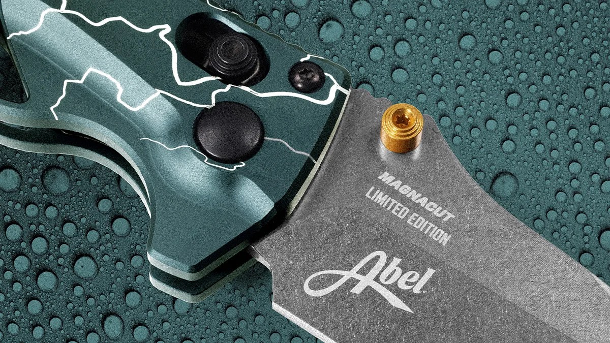 Close-up of a green and silver folding knife blade with "MAGNACUT LIMITED EDITION Abel" text and a gold screw on a water-droplet surface.