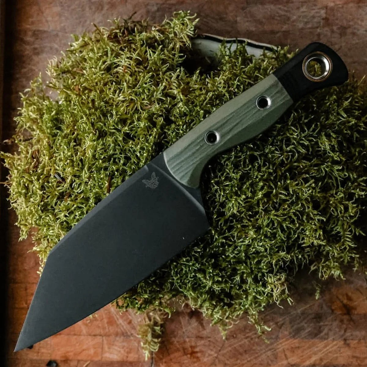 Black wide blade knife with green and black handle resting on green moss on a wooden surface.