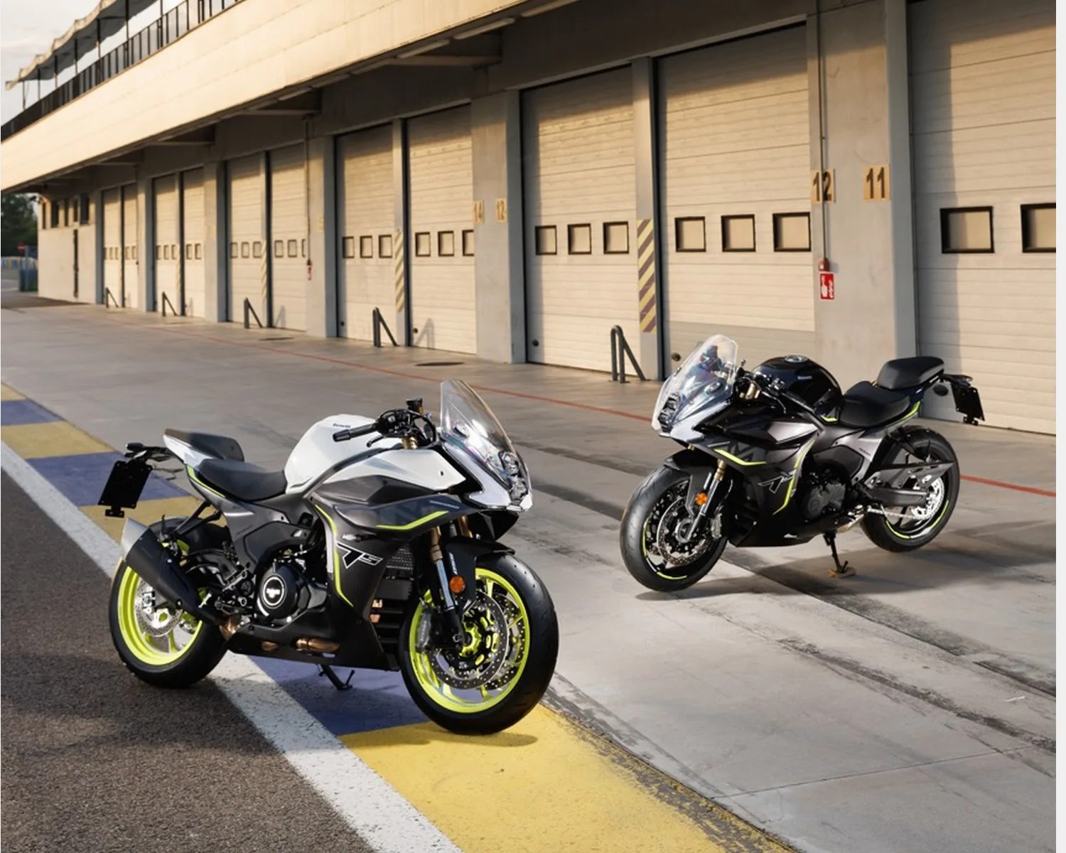 Two sport motorcycles, one white and one black, with neon yellow accents parked on a racetrack pit lane.