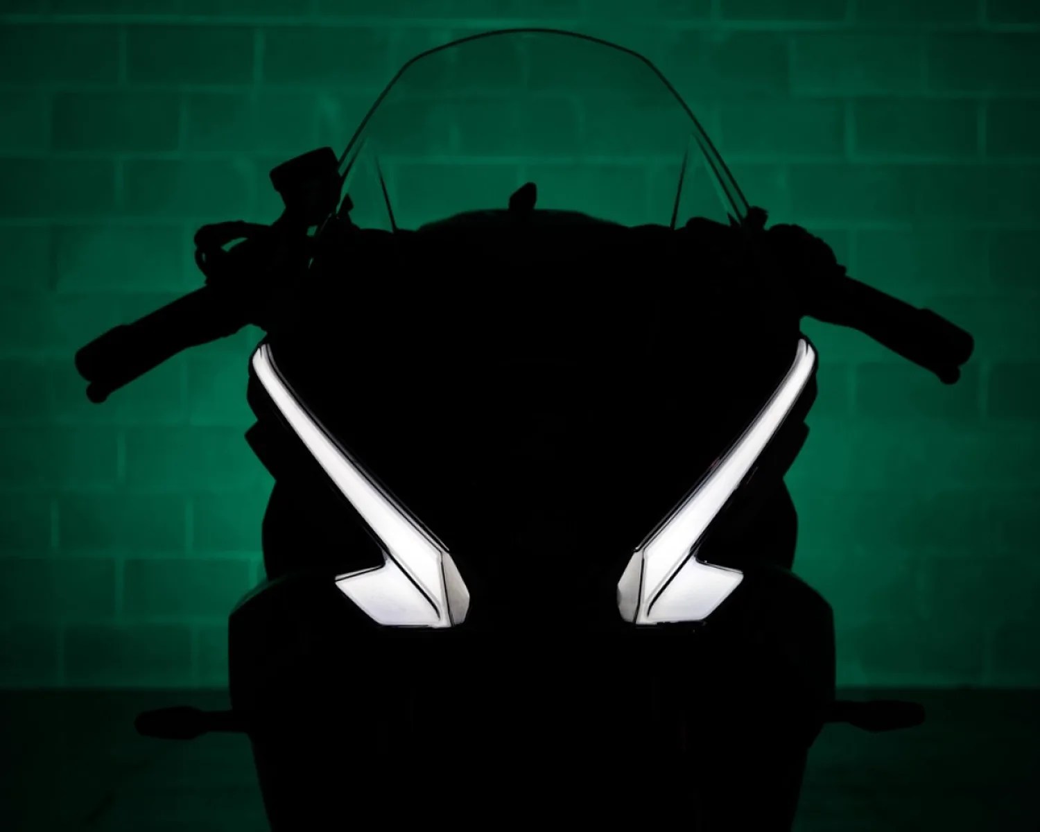 Front view of a motorcycle with illuminated angular LED headlights against a dark green brick wall background.
