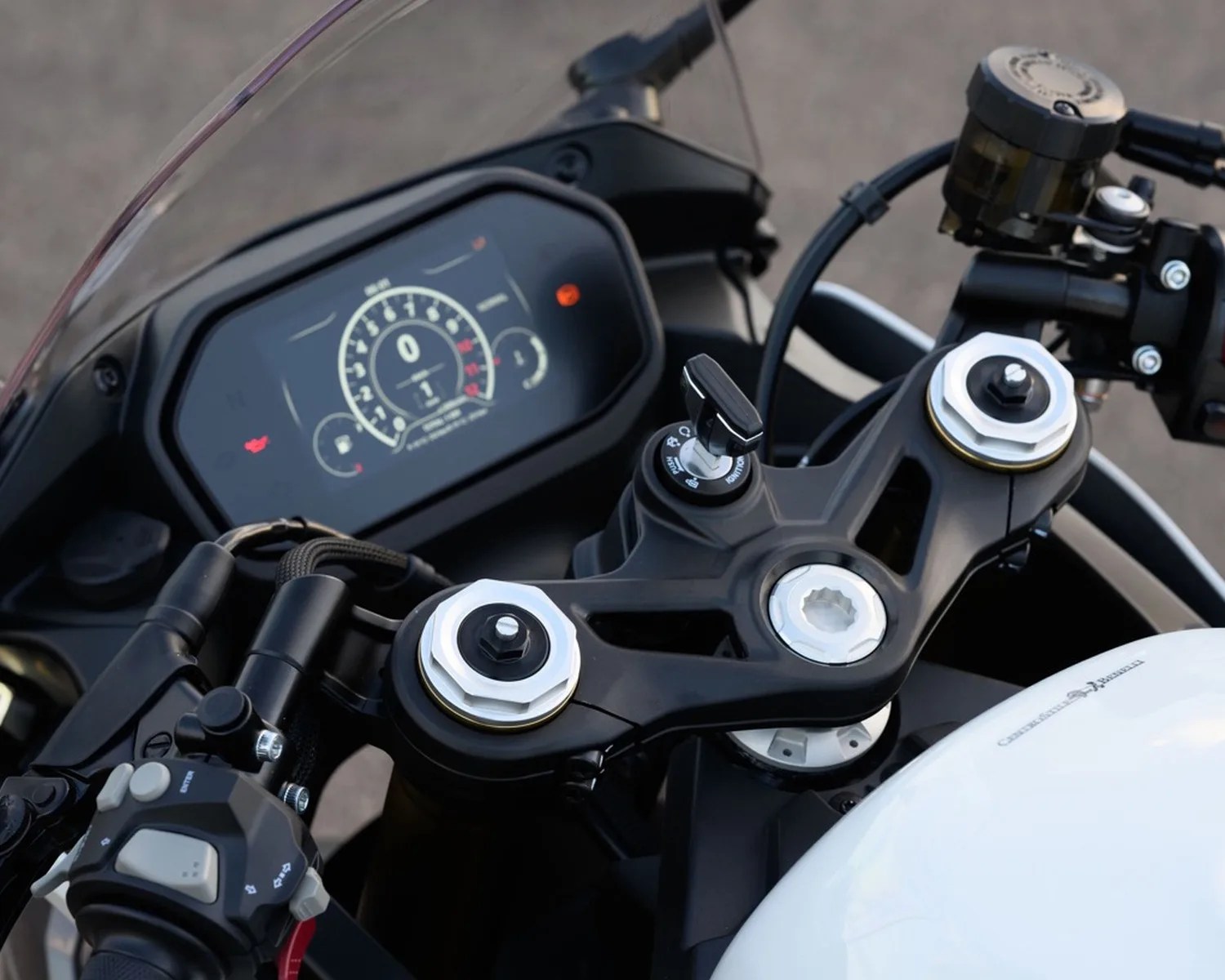 Close-up of a motorcycle dashboard with a digital speedometer, black handlebars, and a white fuel tank.