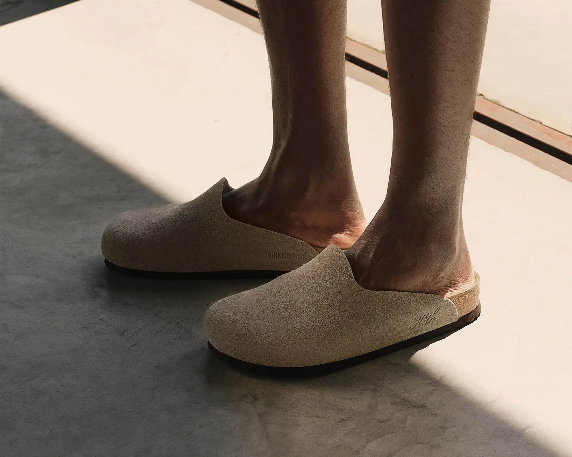 Beige suede slip-on shoes worn on feet standing on a concrete floor with sunlight and shadow.