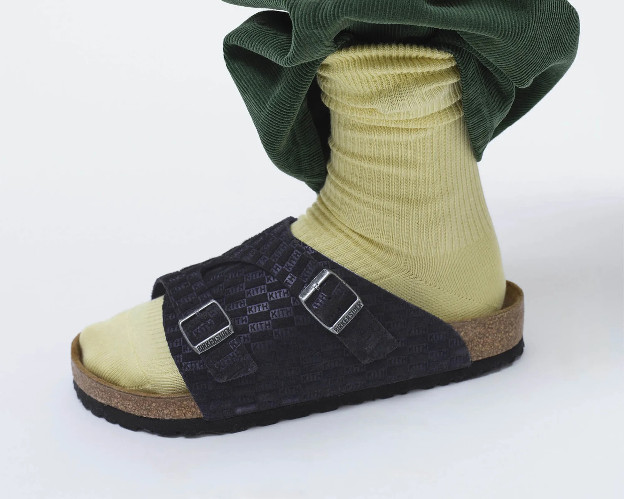 Black Birkenstock sandal with KITH logo pattern and silver buckles worn with light yellow ribbed socks and green pants.