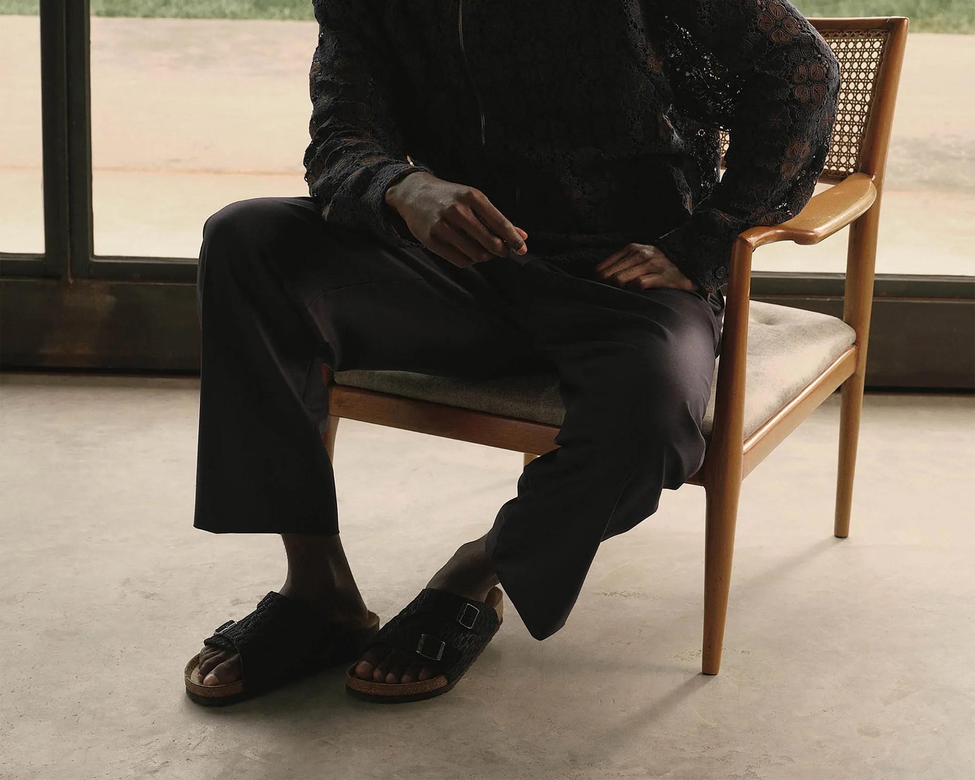 Person wearing black wide-leg pants and black sandals sitting on a wooden chair with a woven backrest.
