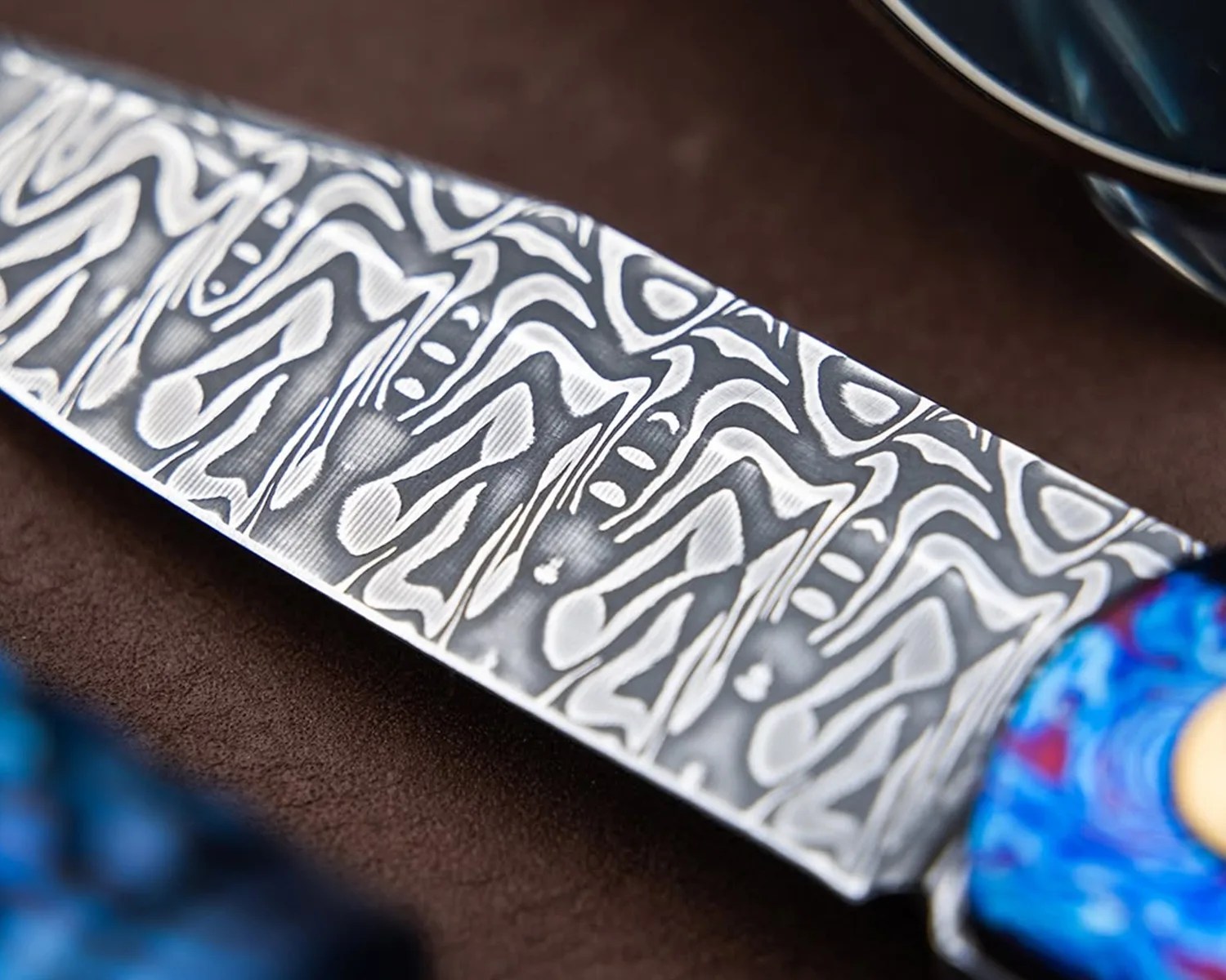Close-up of a knife blade with intricate Damascus steel pattern and a blue handle.