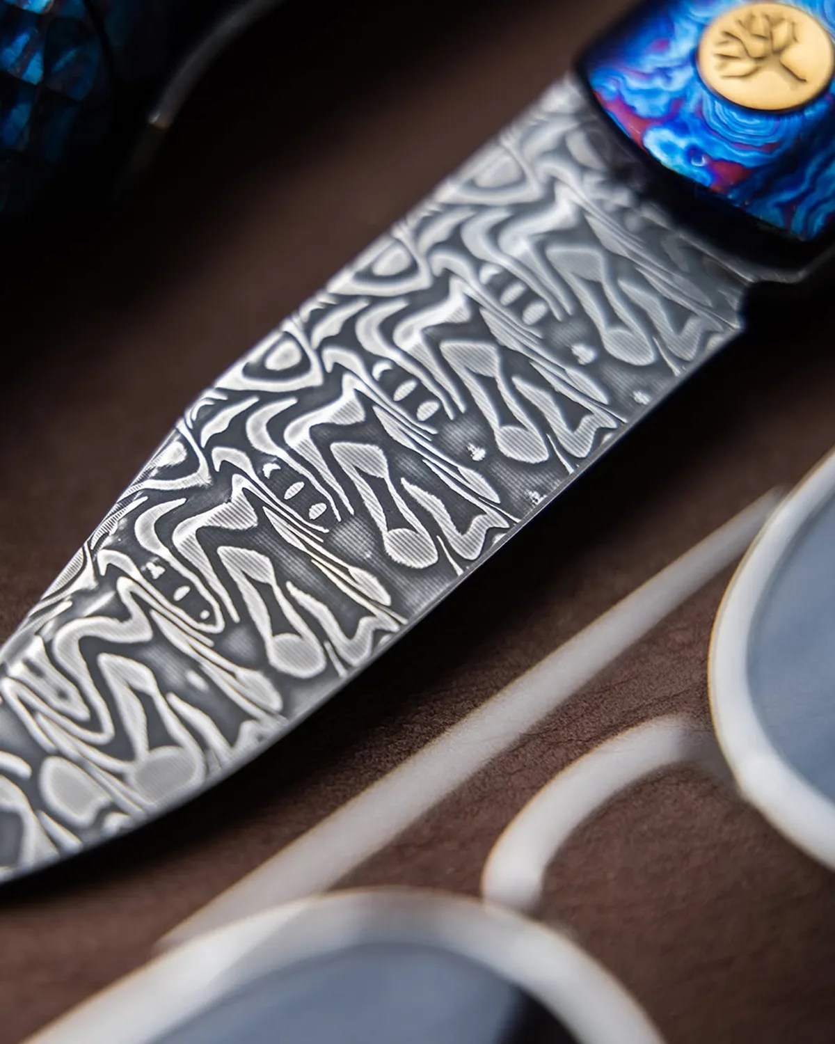 Close-up of a Damascus steel knife blade with intricate wave patterns and a blue handle featuring a gold tree emblem.