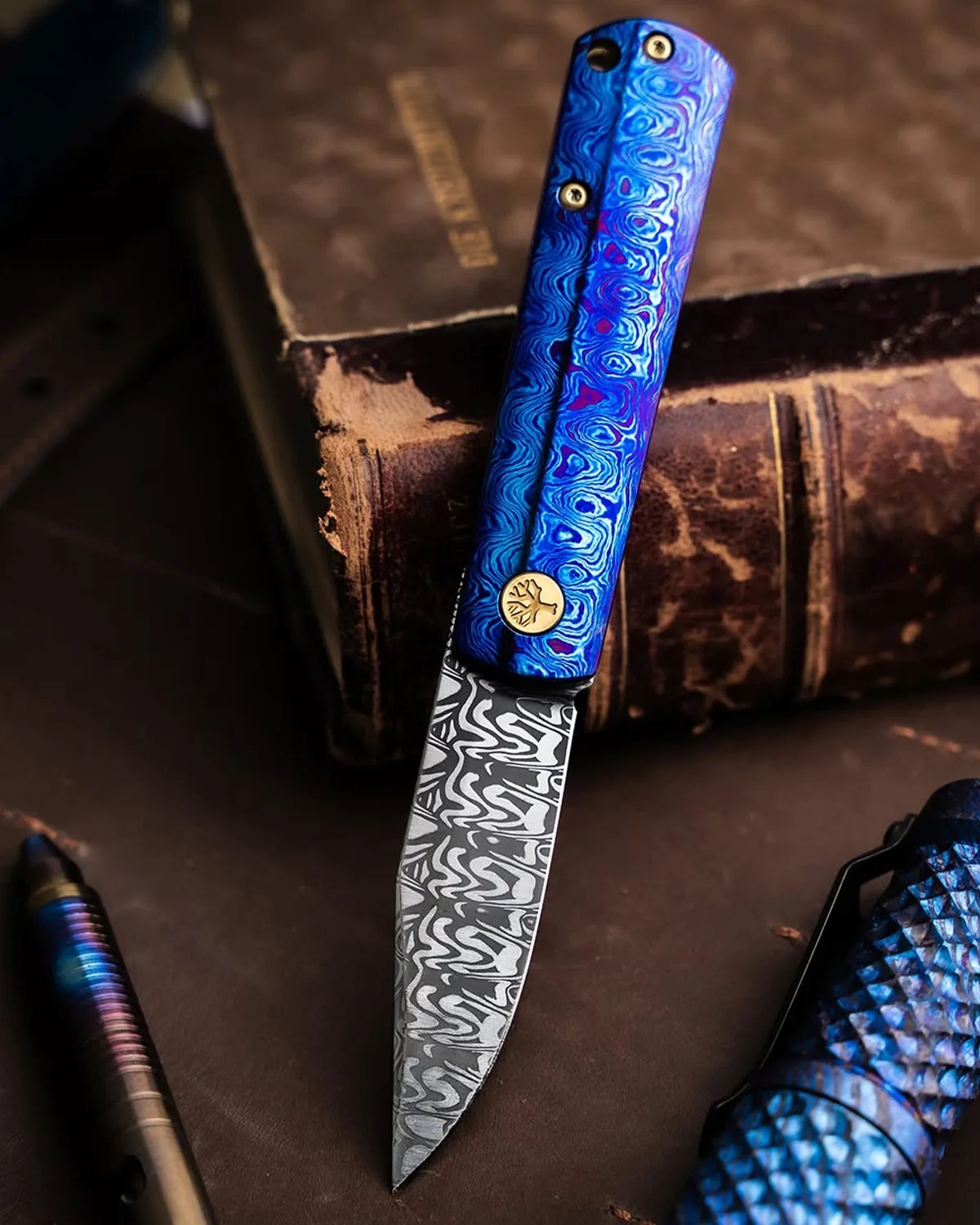 Folding knife with a blue patterned handle and a Damascus steel blade resting on a worn brown book.