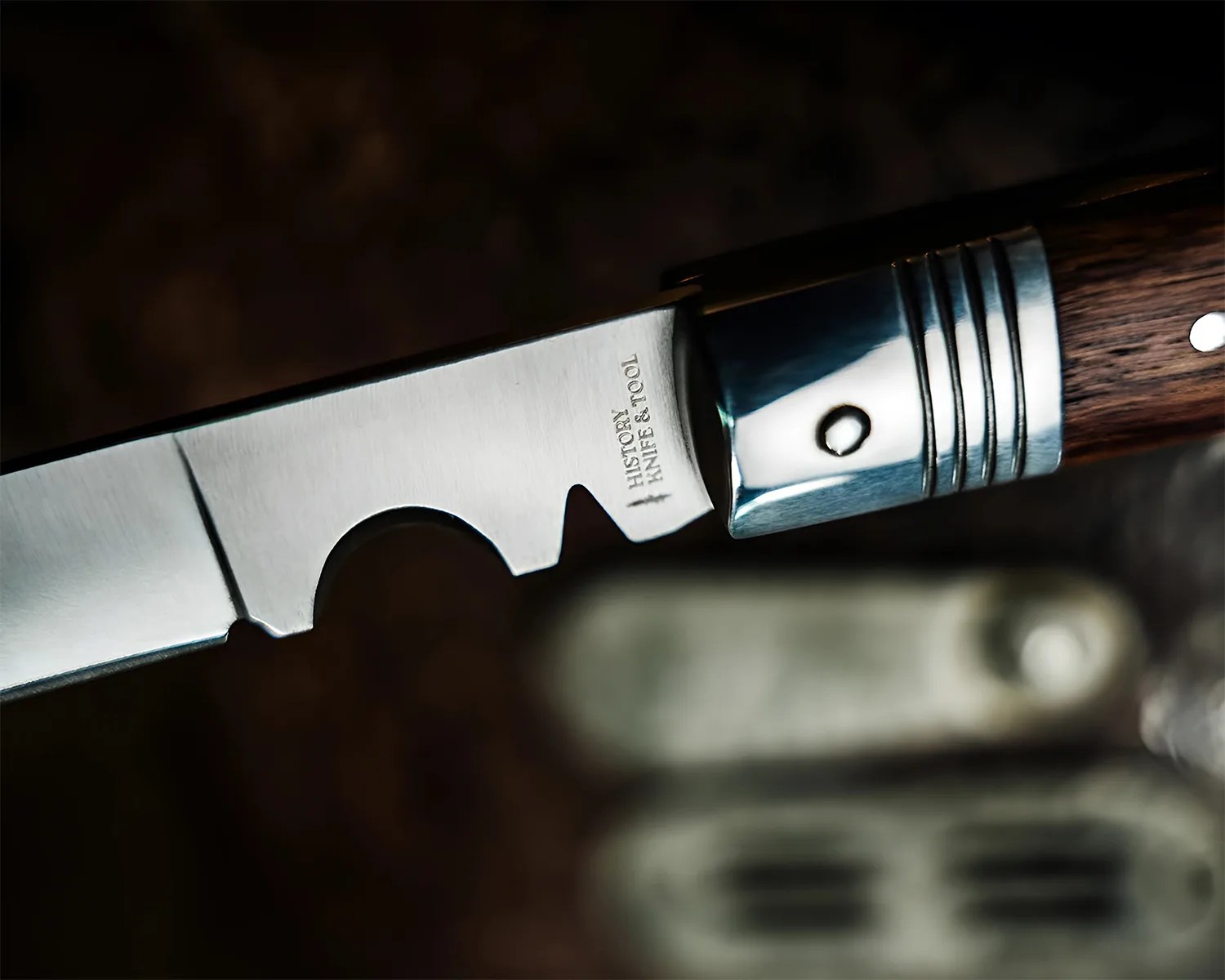 Close-up of a folding knife blade with "HISTORY KNIFE & TOOL" engraved near the wooden handle with metal rivets.