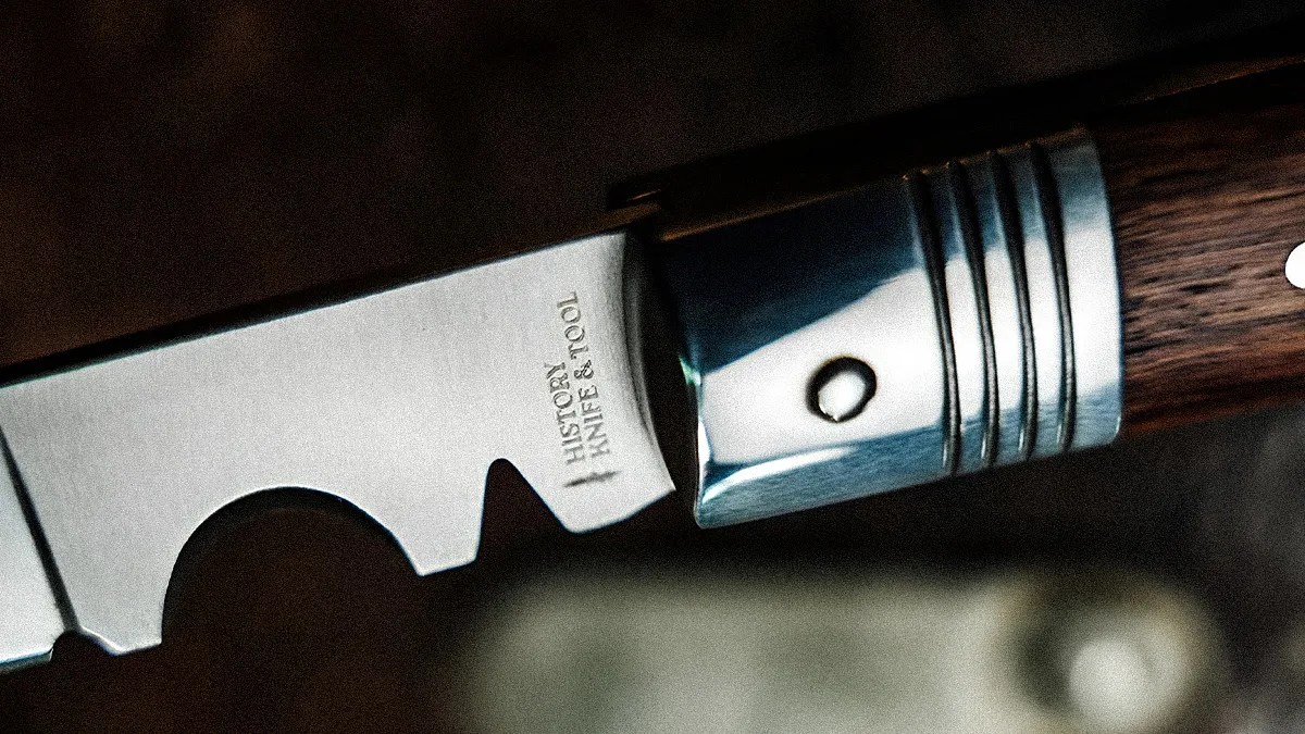 Close-up of a serrated knife blade with "HISTOR KNIFE & TOOL" engraved near a polished metal bolster and wooden handle.