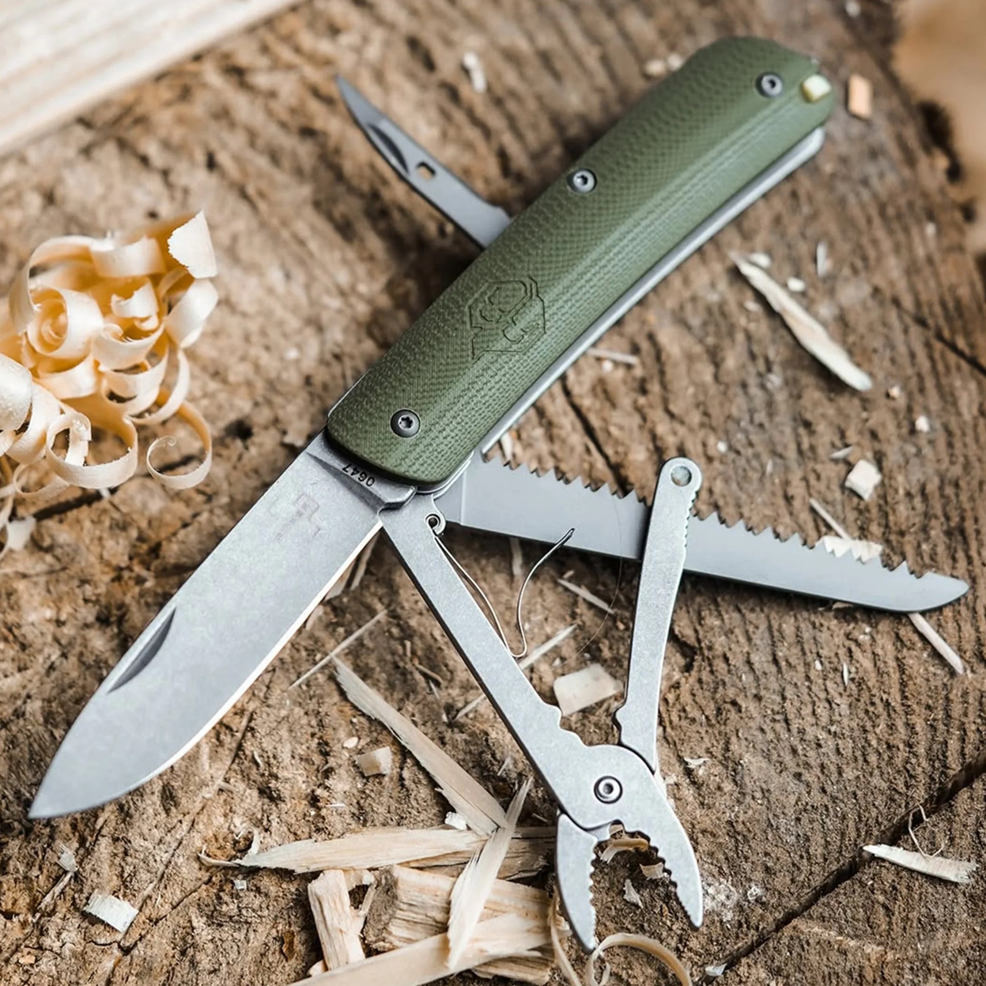 Multi-tool with green textured handle and various metal tools extended, resting on wood shavings and dirt.