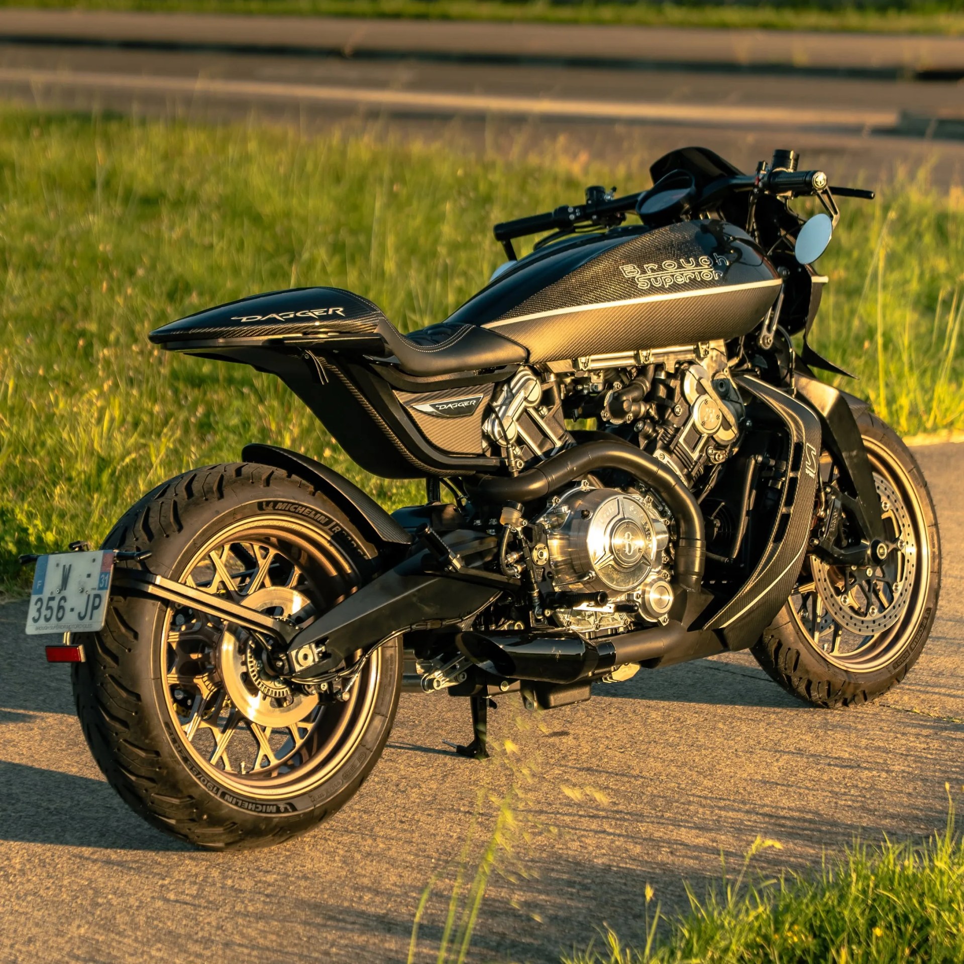 Black Brough Superior Dagger motorcycle with carbon fiber finish and gold rims parked on pavement near grass.