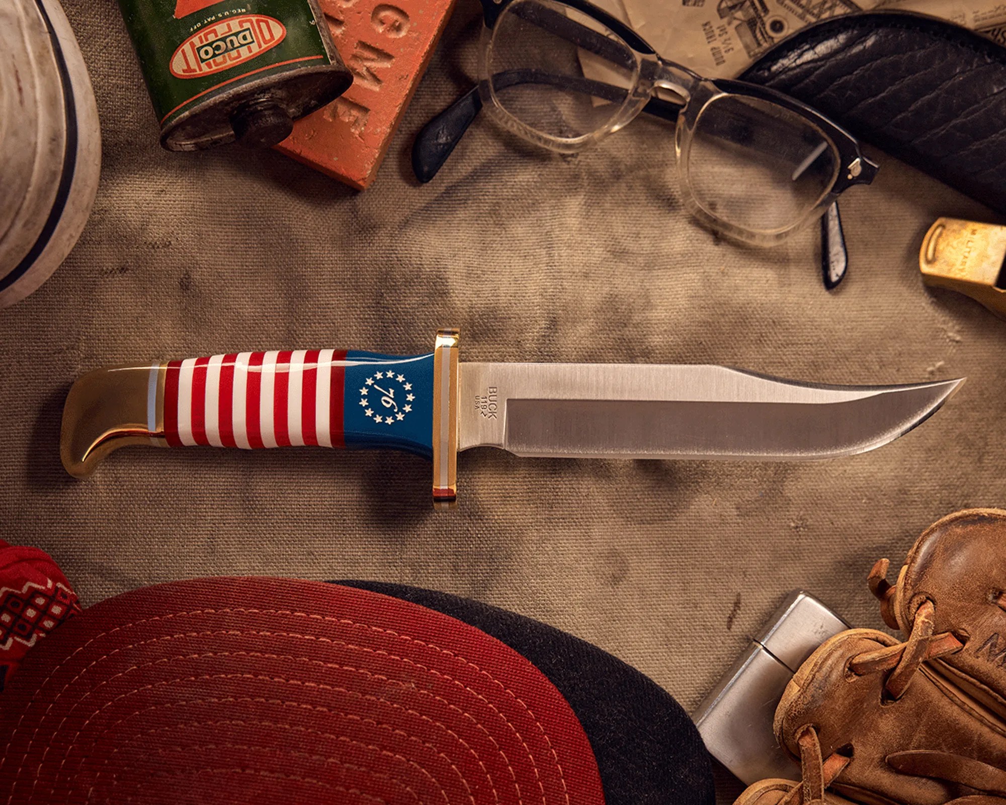 Knife with American flag handle and brass guard on a fabric surface near glasses, a red cap, and a leather glove.