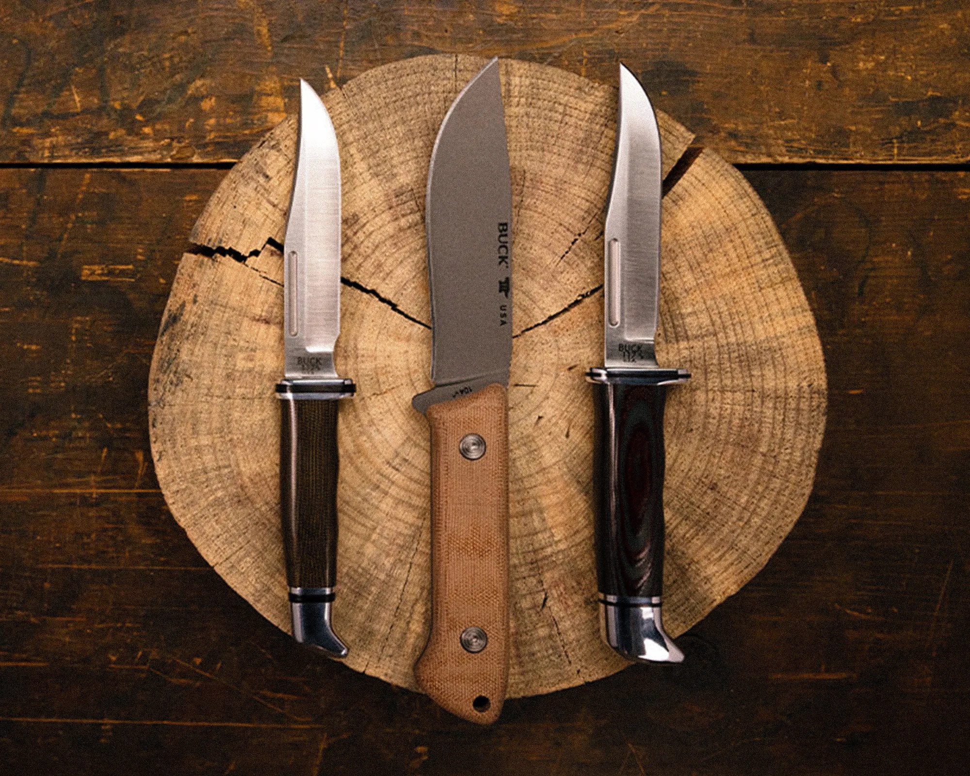 Three fixed-blade knives with different handle colors arranged on a round wooden log slice on a dark wooden surface.