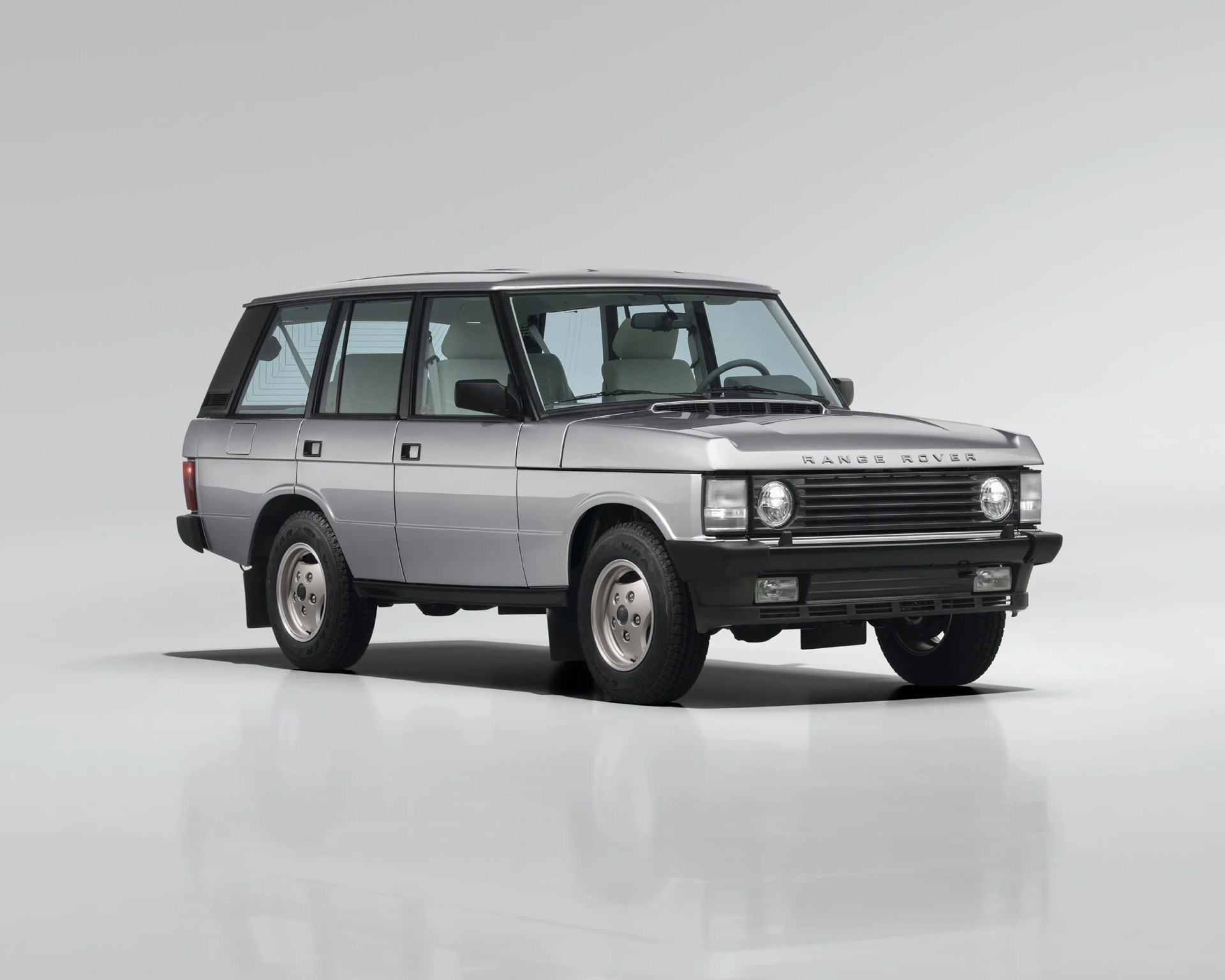 Silver classic Range Rover SUV with black trim and white interior seats on a white reflective surface.