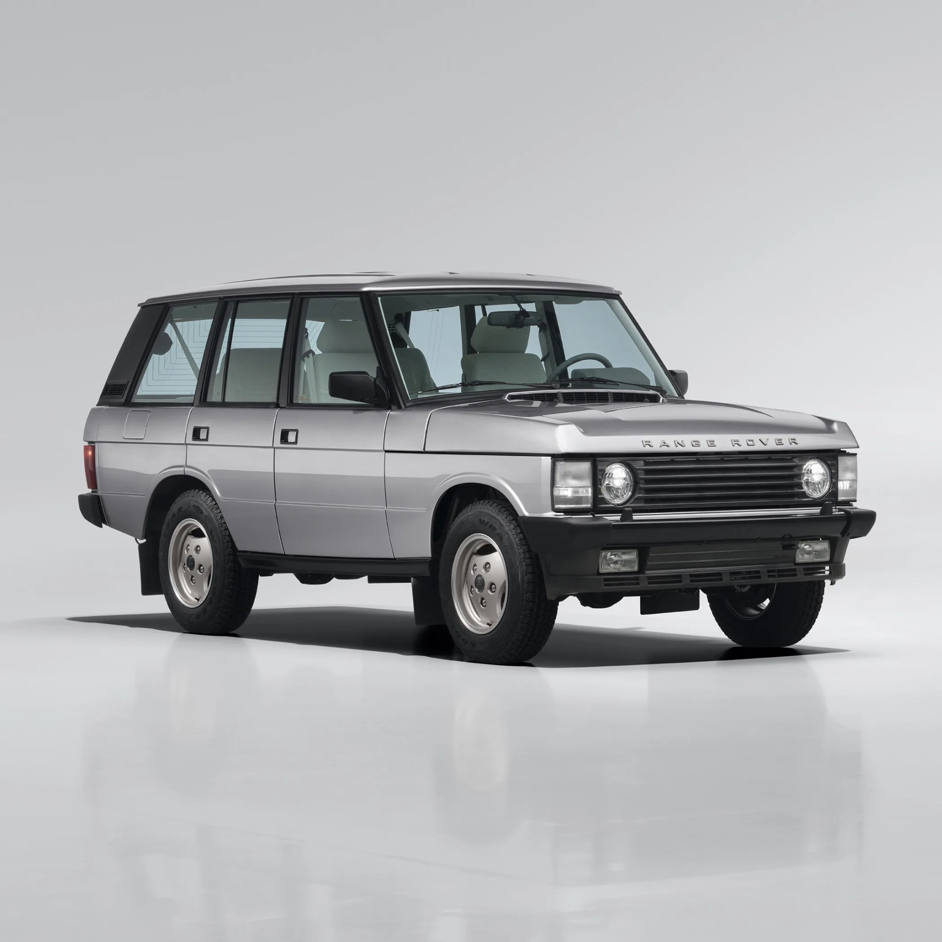 Silver classic Range Rover SUV with black trim and white interior seats on a white reflective floor.