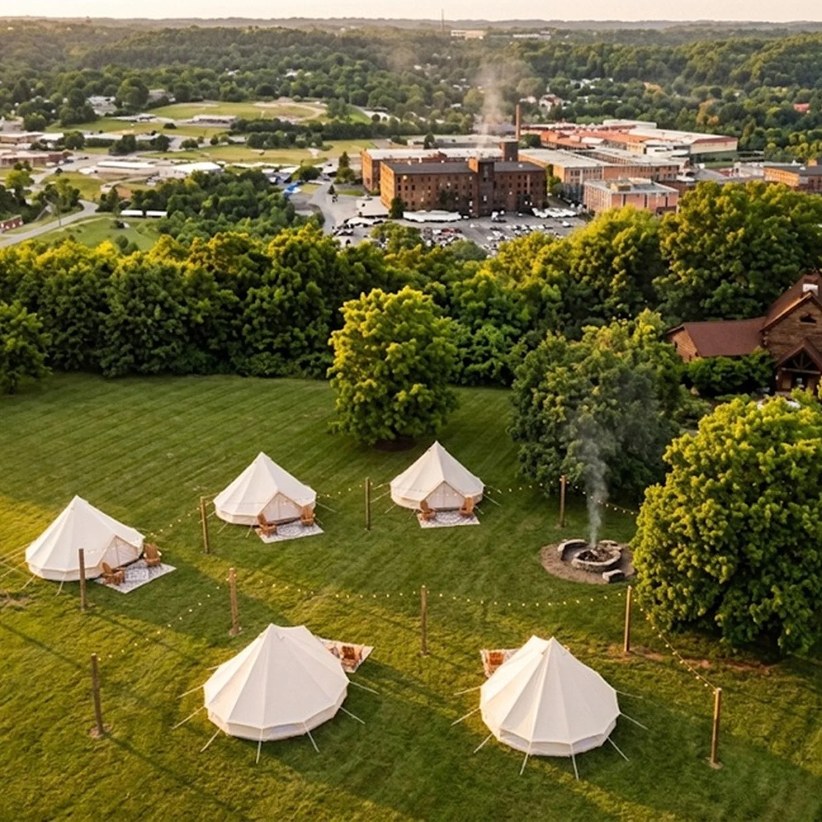 White canvas tents arranged on a grassy field with a smoking fire pit and dense green trees in the background.