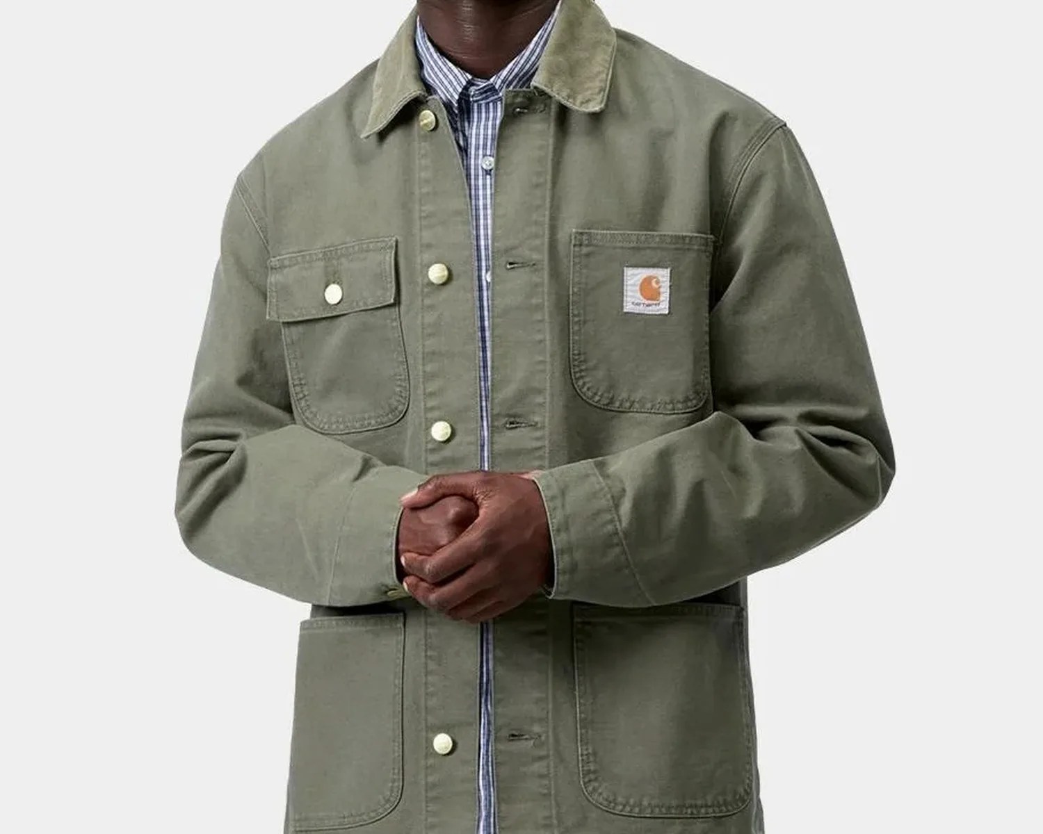 Olive green Carhartt work jacket with buttoned flap and patch pockets worn over a blue striped shirt.
