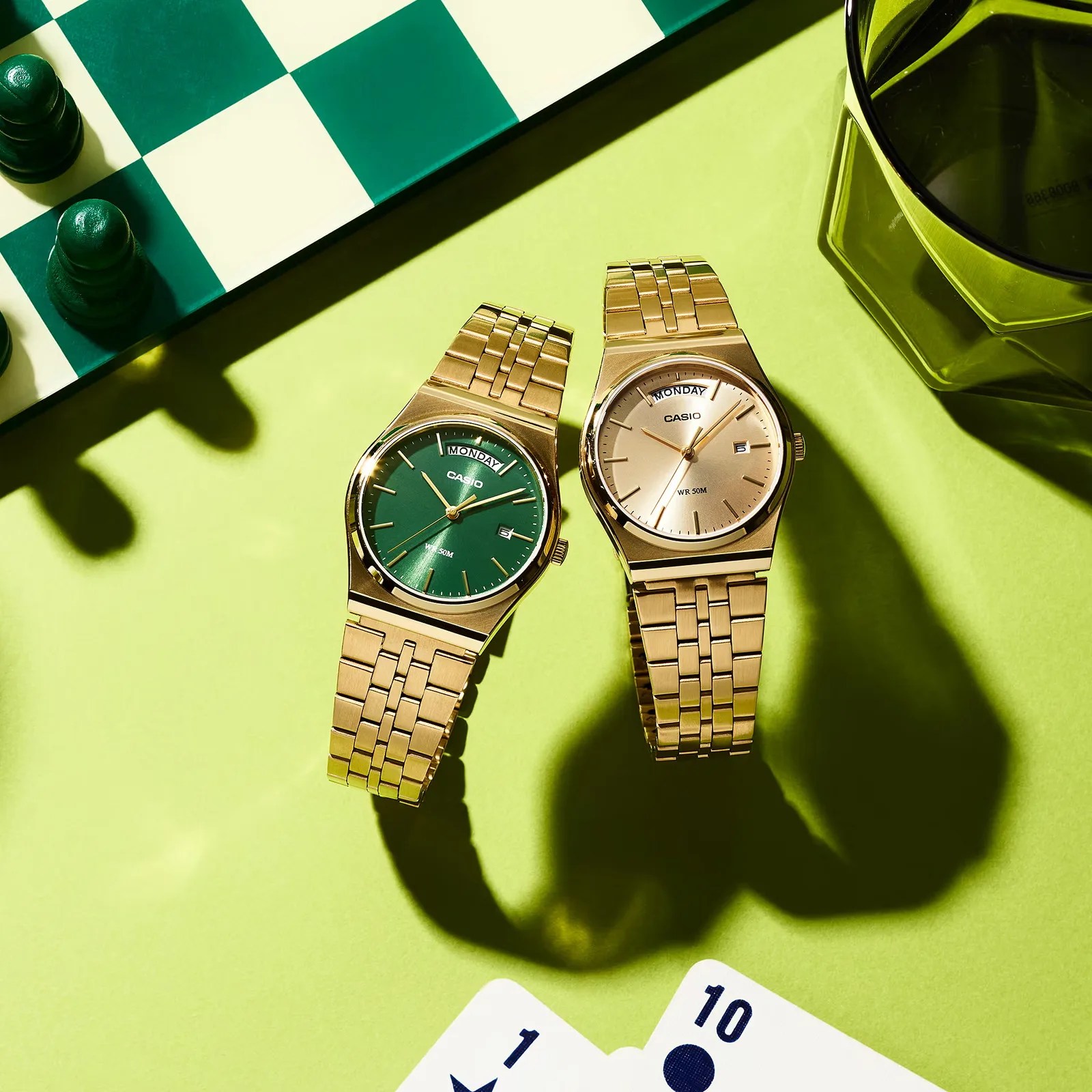 Two gold Casio wristwatches with green and gold faces on a green surface near a chessboard and playing cards.