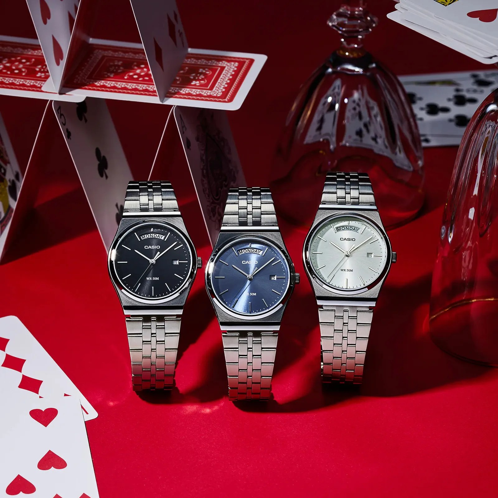 Three Casio wristwatches with silver metal bands and black, blue, and white faces on a red surface with playing cards and upside-down glasses.