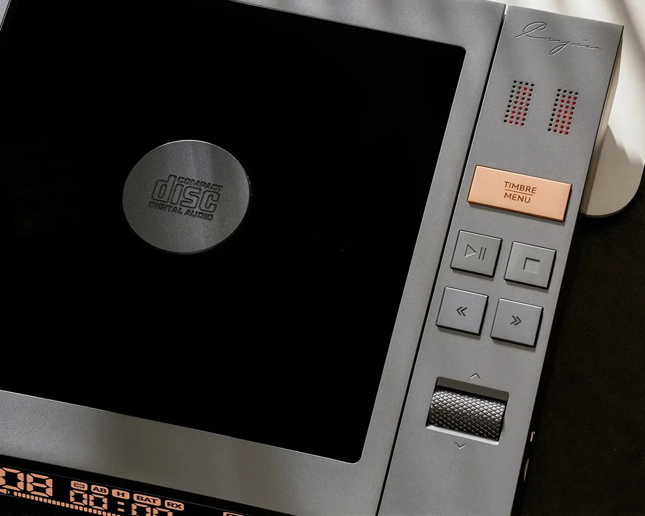 Close-up of a gray compact disc player with a black display, playback control buttons, a textured volume knob, and a copper-colored "TIMBRE MENU" button.