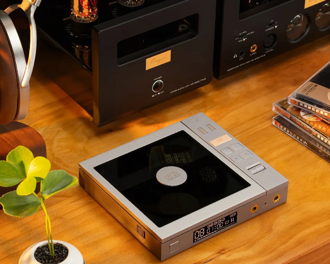 Silver portable CD player with black top and digital display on wooden table near headphones and stacked CDs.