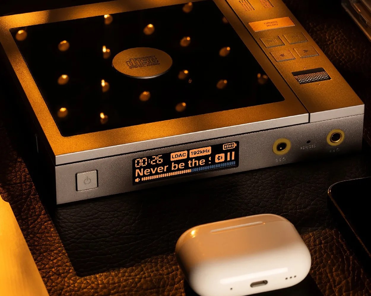 Portable digital audio player with a small screen displaying "Never be the" and a white wireless earbud case nearby.