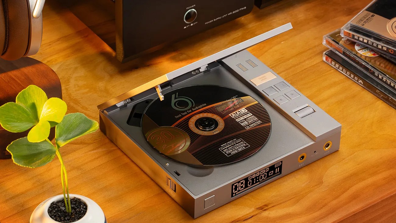 Silver portable CD player with a black CD inside on a wooden surface next to a small potted plant and stacked CDs.