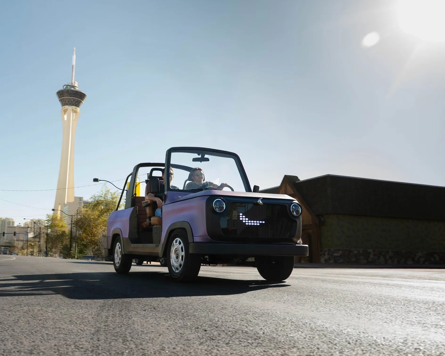 Purple electric convertible car with two passengers driving on a sunny street near a tall observation tower.