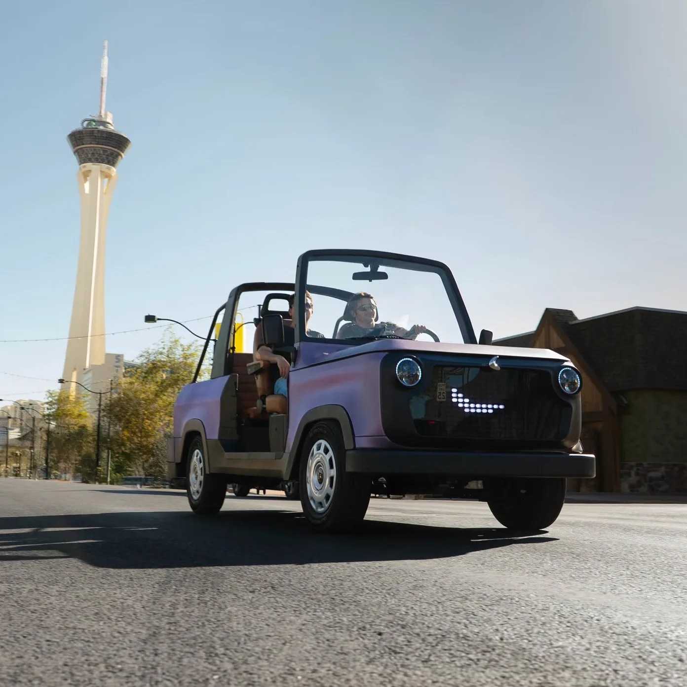 Purple compact electric convertible car with digital smiley face grille driving on a city street.