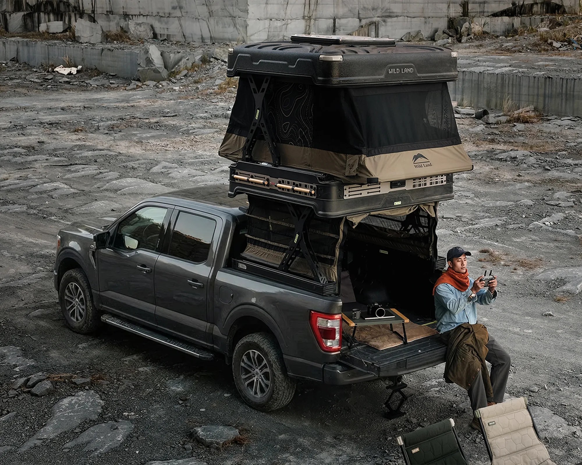 Gray pickup truck with a rooftop tent setup and a person sitting on the tailgate holding a remote controller.