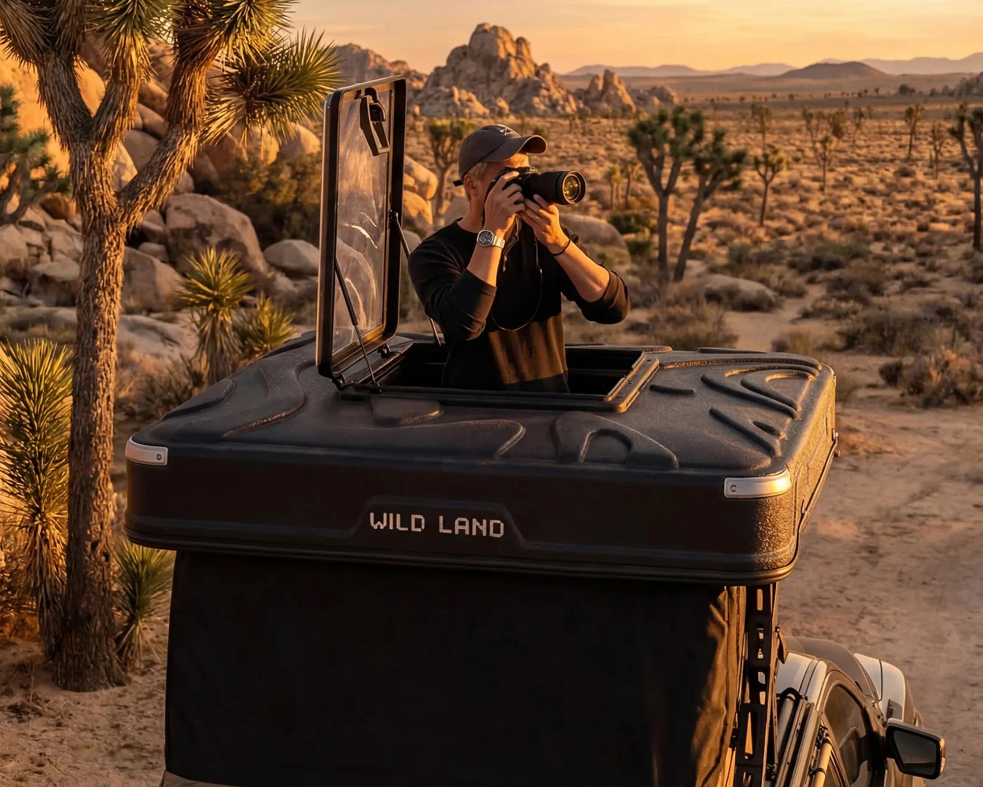 Person using a camera while standing inside a rooftop tent labeled "WILD LAND" on a vehicle in a desert landscape.