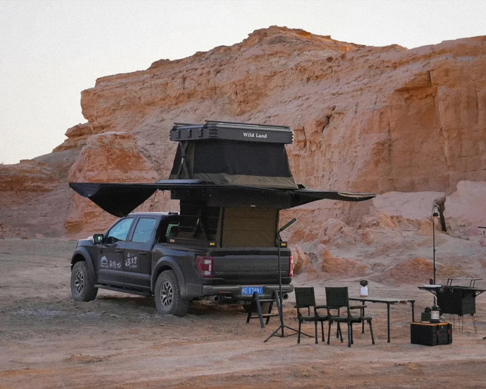 Black pickup truck with rooftop tent labeled "Wild Land" parked in a desert with camping chairs and table nearby.