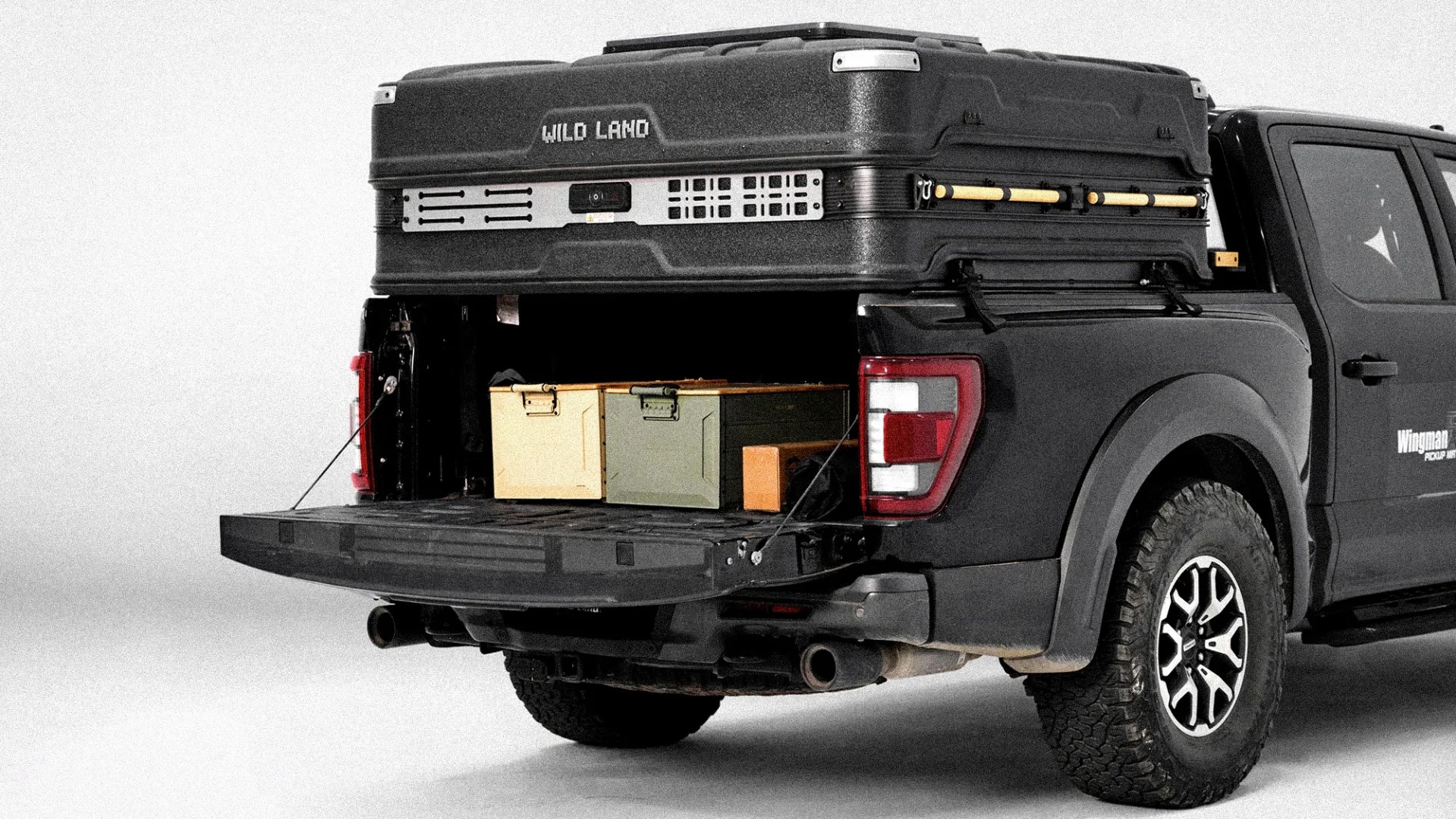 Black pickup truck with open tailgate carrying two metal storage boxes and a large black rooftop cargo box labeled "WILD LAND.