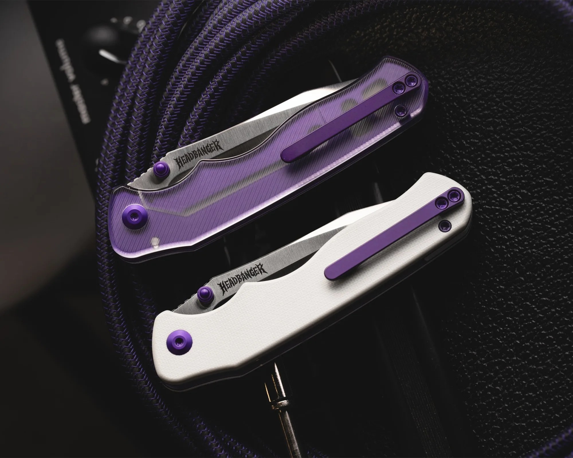 Two folding knives with "Headbanger" engraved on the blades, one with a purple translucent handle and the other with a white handle, both with purple clips.