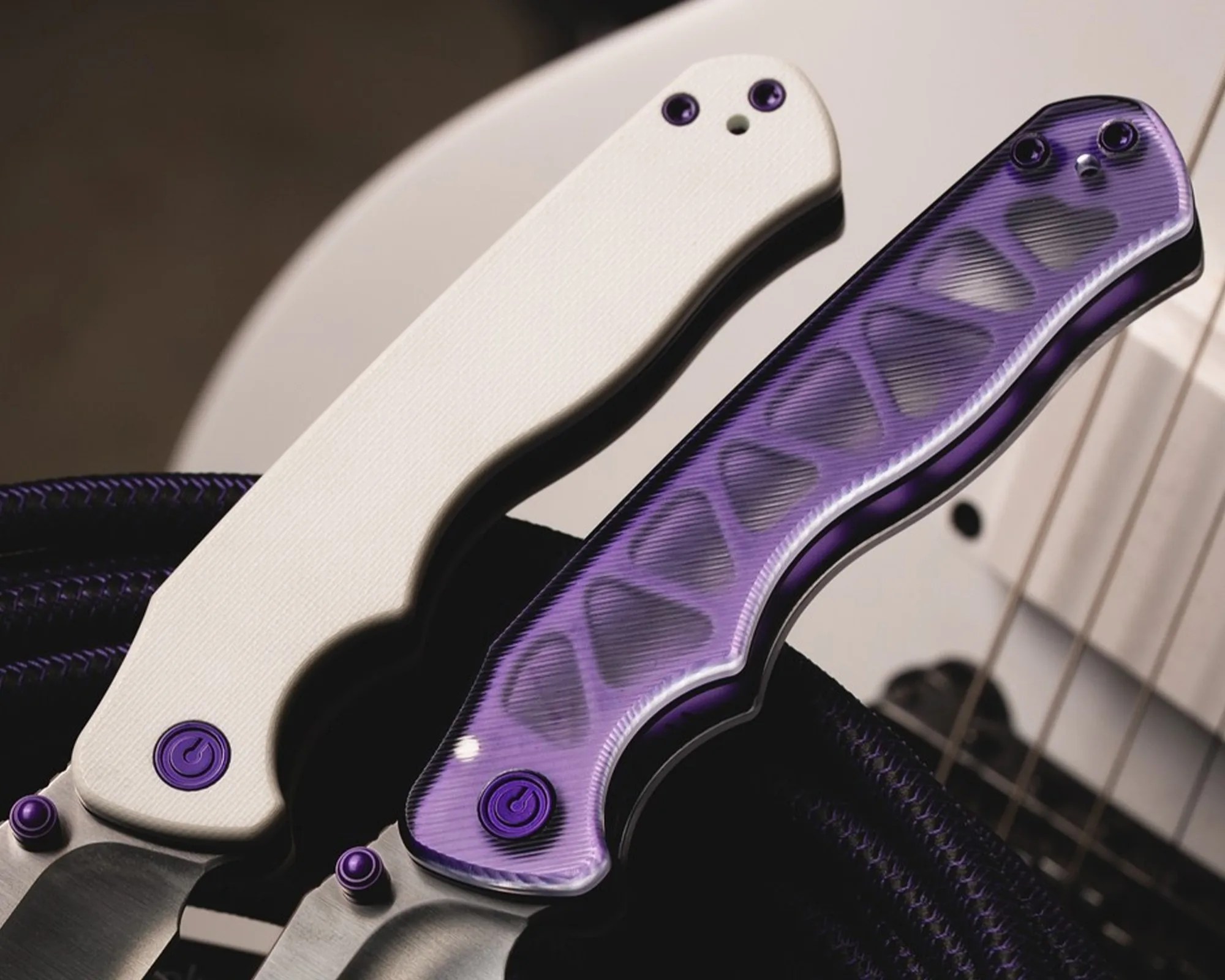 Two folding knives with textured handles, one white and one translucent purple, resting on a guitar body.