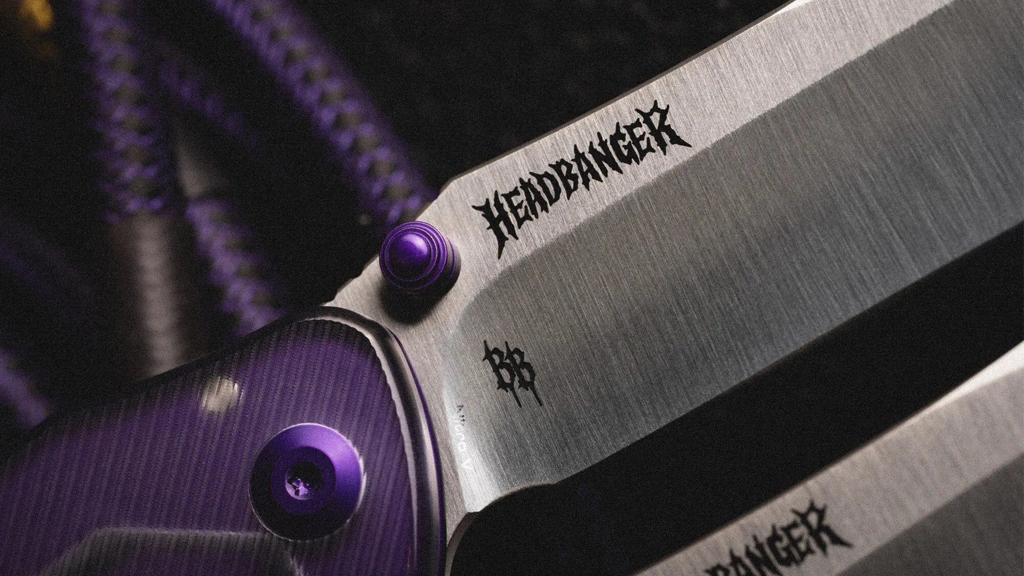 Close-up of a folding knife blade with "HEADBANGER" engraved and purple handle screws.