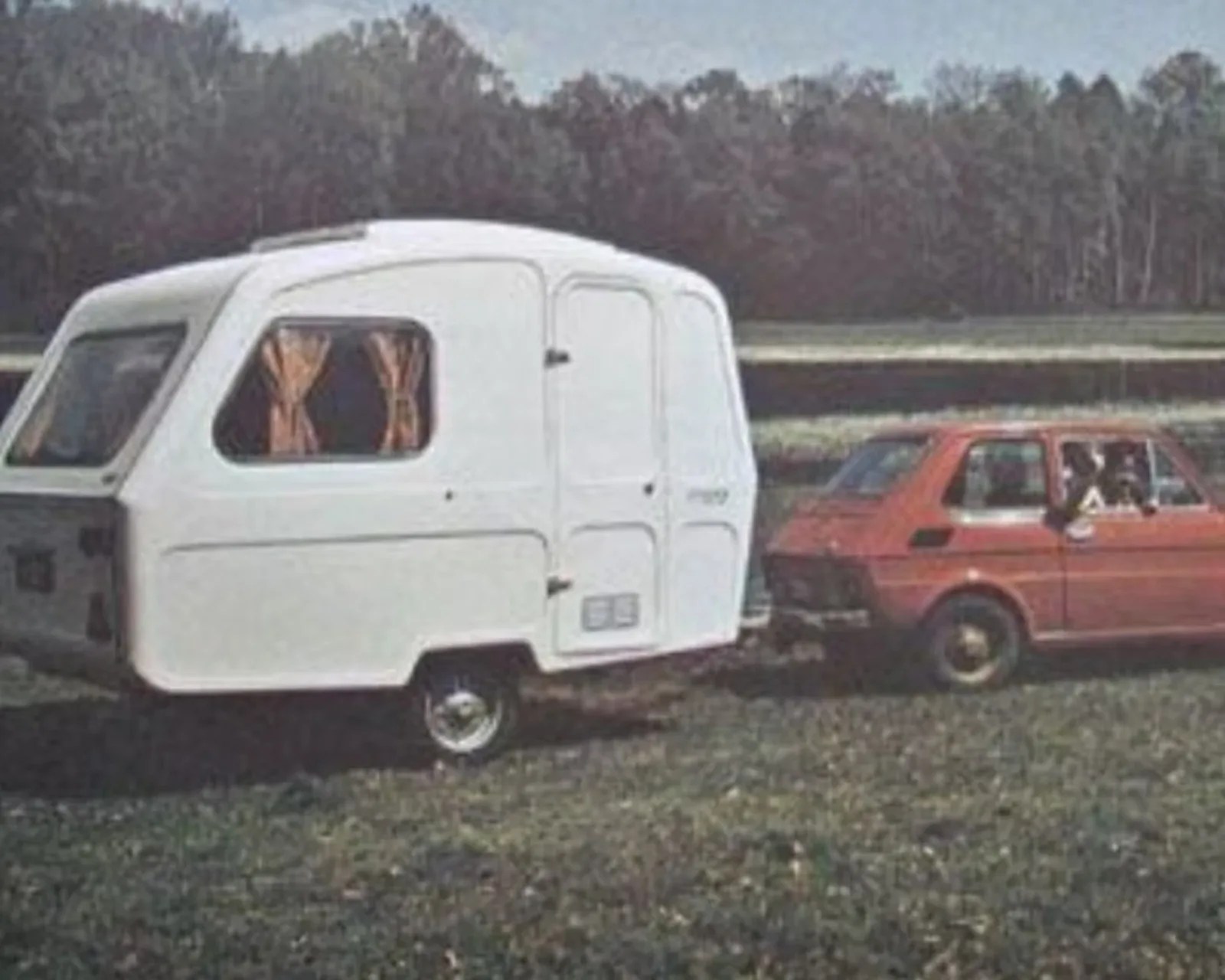 Red compact car towing a small white caravan with orange curtains in a grassy field.