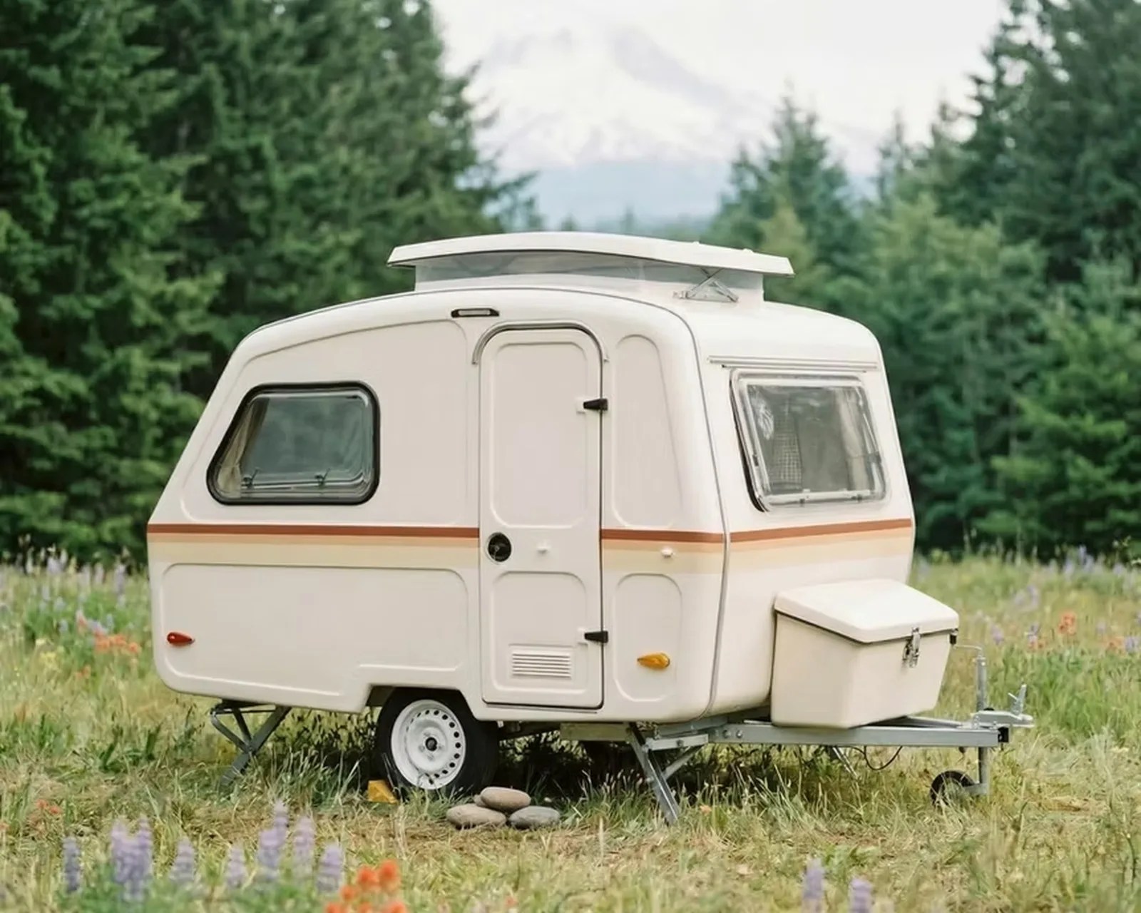 Compact white travel trailer with orange stripe and pop-up roof parked in a grassy field with trees.