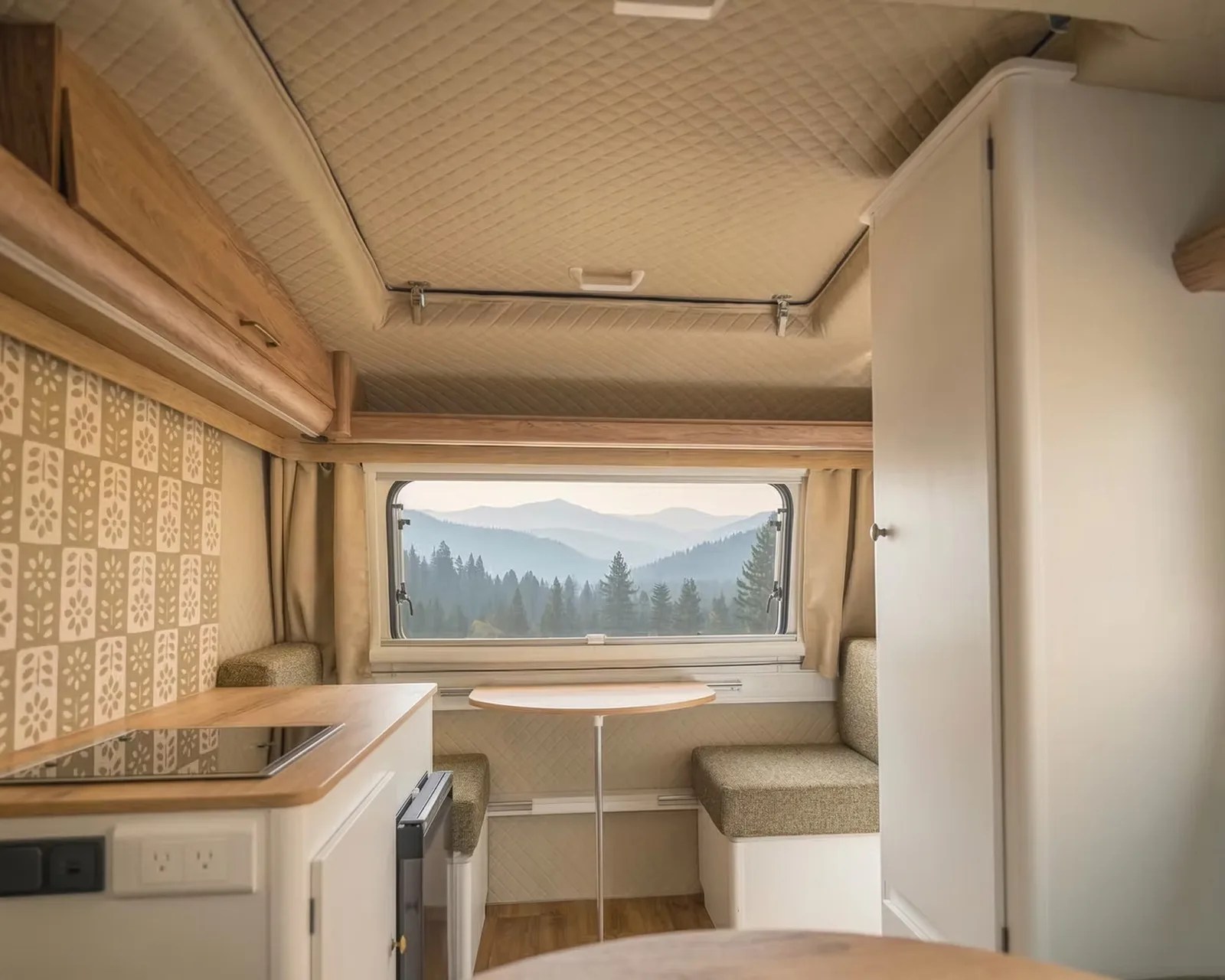 Interior of a beige and wood-finished camper with a small table, cushioned bench seats, and a window showing a mountain view.