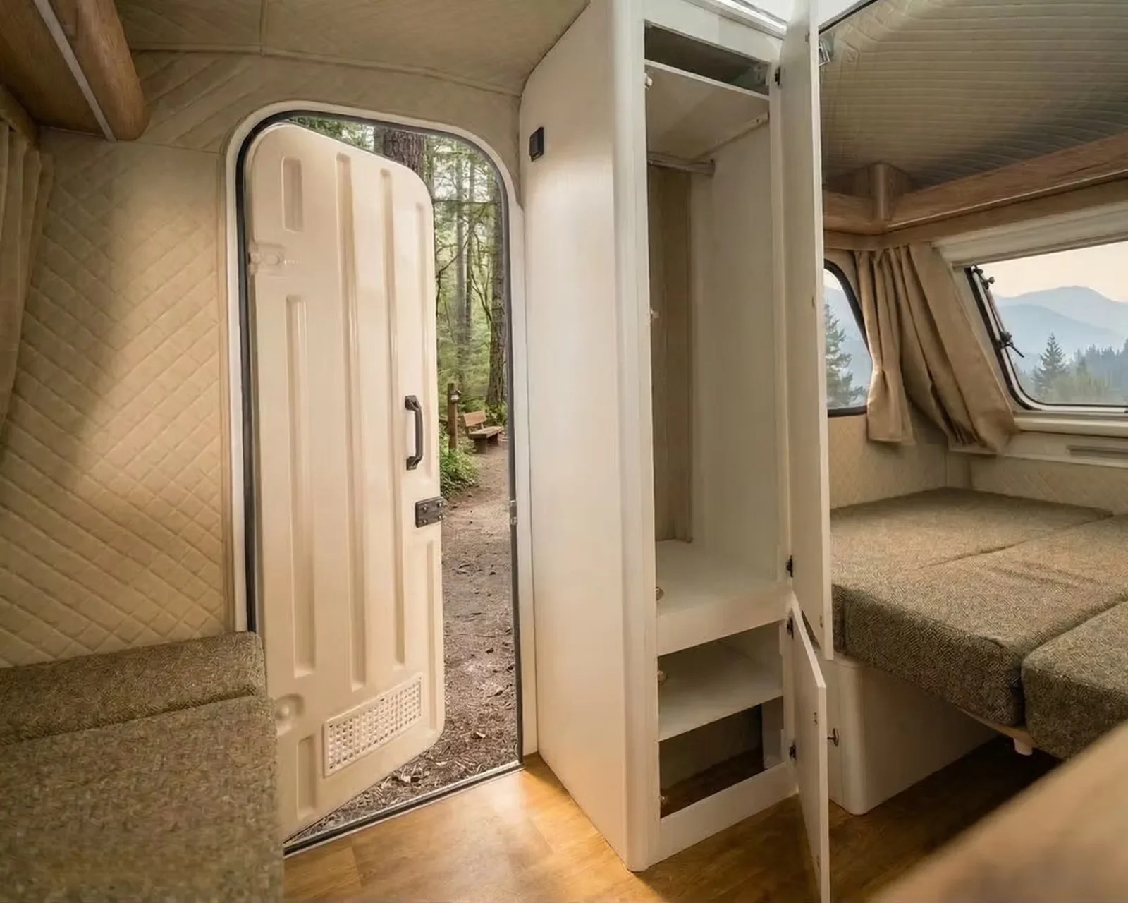 Interior of a camper van showing an open beige door, empty white wardrobe, and a bed with brown cushions near windows with beige curtains.