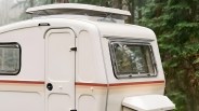 White camper trailer with a pop-up roof vent and horizontal red and beige stripes parked near green trees.