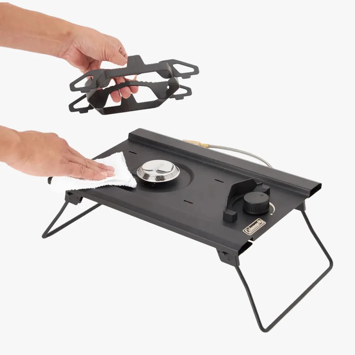 Black Coleman portable camping stove with foldable legs, burner, control knob, and a hand holding a cleaning cloth and a detachable grate.